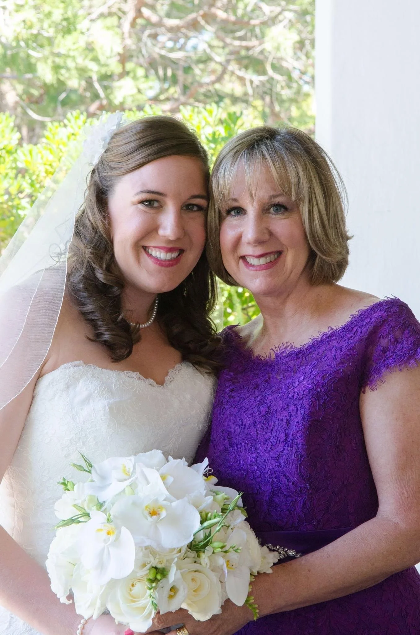 A bride in a white wedding dress holding a bouquet of white flowers poses with a woman wearing a purple dress. They are smiling and standing outside with greenery in the background.