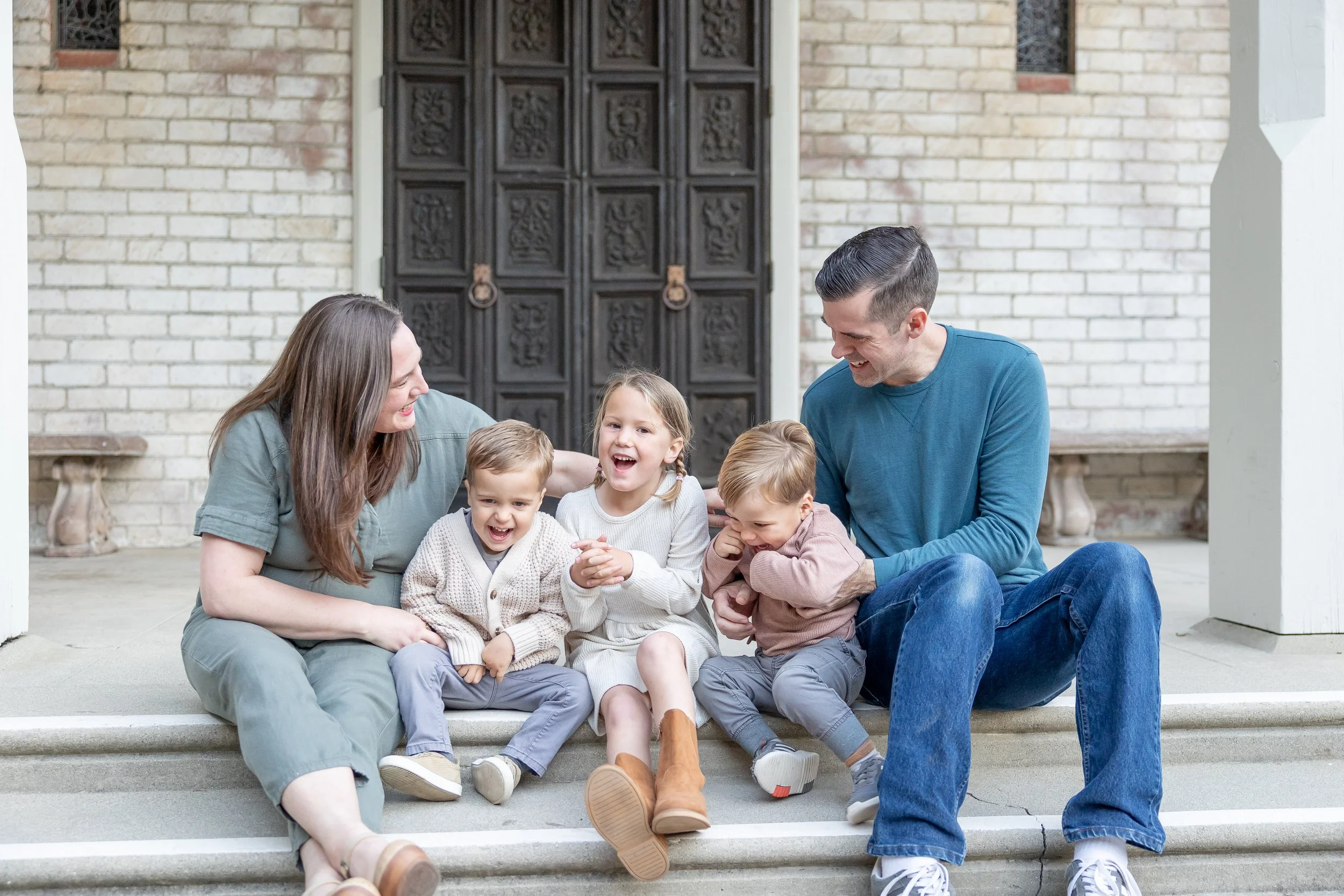 A family with two adults and three children sitting on steps in front of a brick wall and a large wooden door, smiling and enjoying each other's company.