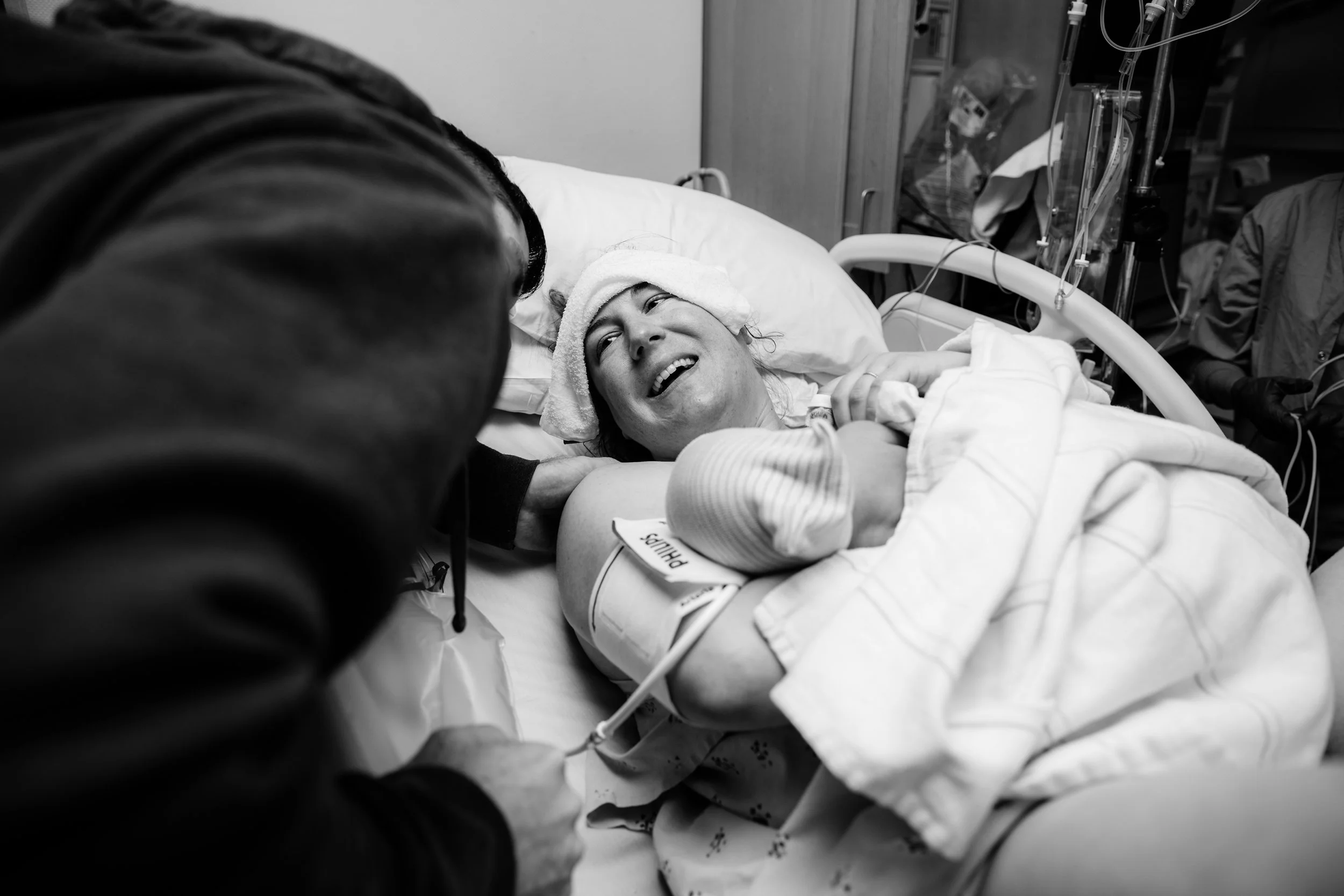 A woman in a hospital bed smiles at a visitor who is leaning over her. The woman has a towel on her head and medical tubes attached to her. The scene appears to be in a hospital room with medical equipment visible in the background.