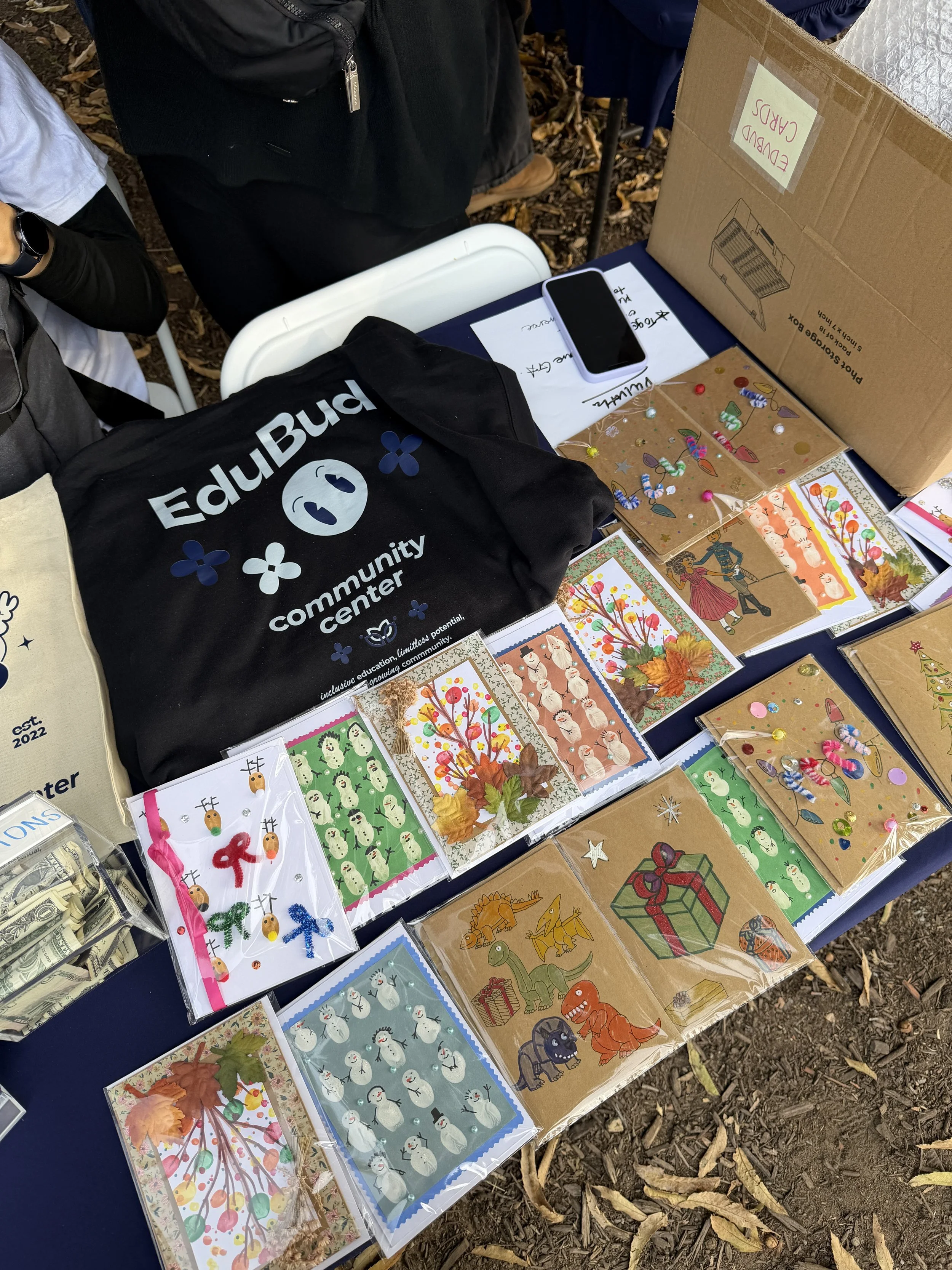 Table with various greeting cards featuring colorful illustrations of snowmen, owls, trees, animals, and holiday themes, along with a black bag labeled 'Edwards Community Center,' a smartphone, and a cardboard box labeled 'Edubd Cards.'