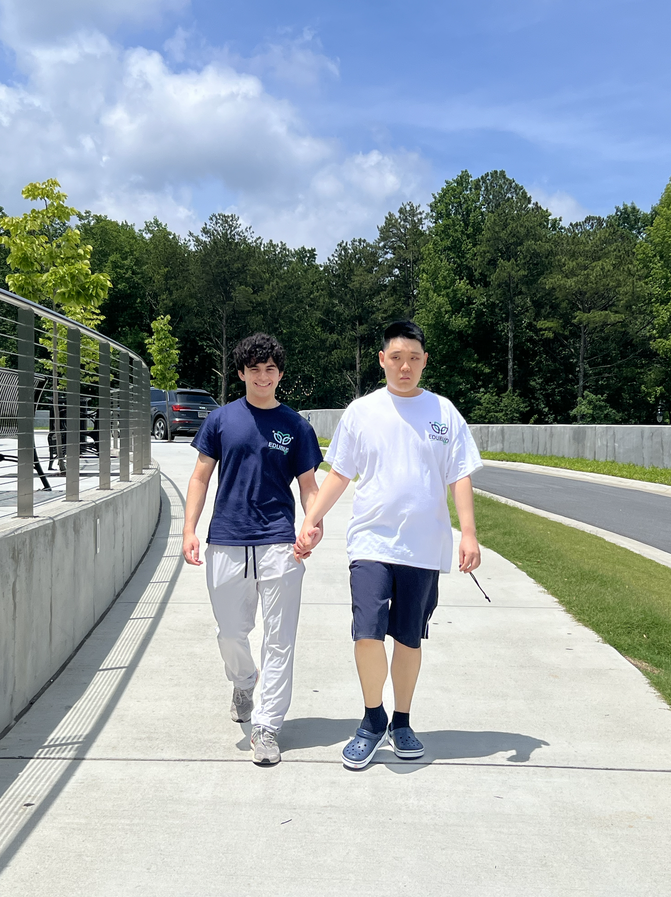 Two young men walking hand in hand outdoors on a sunny day, with trees and a blue sky in the background.