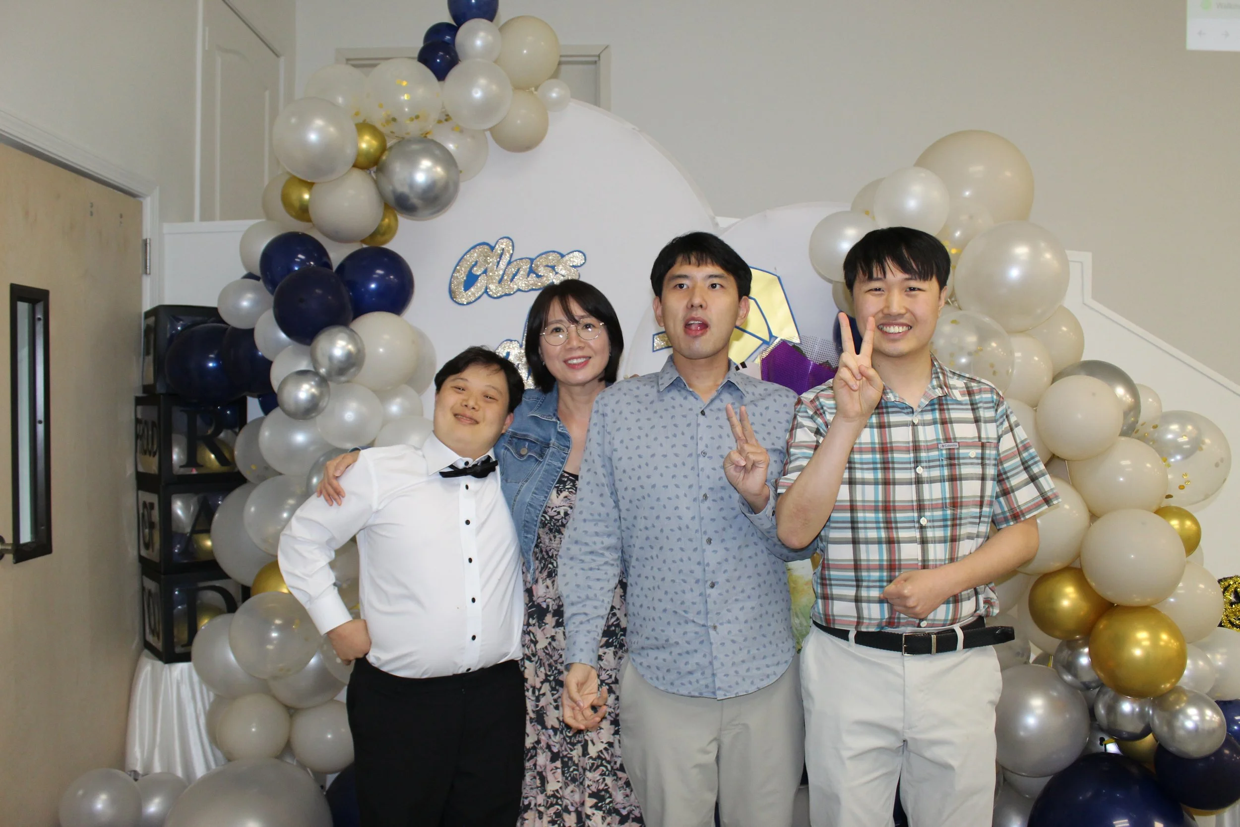 Group of four people standing in front of a balloon decoration with a sign that says "Class". The group includes two men, a woman, and a young boy. They are smiling and making peace signs.