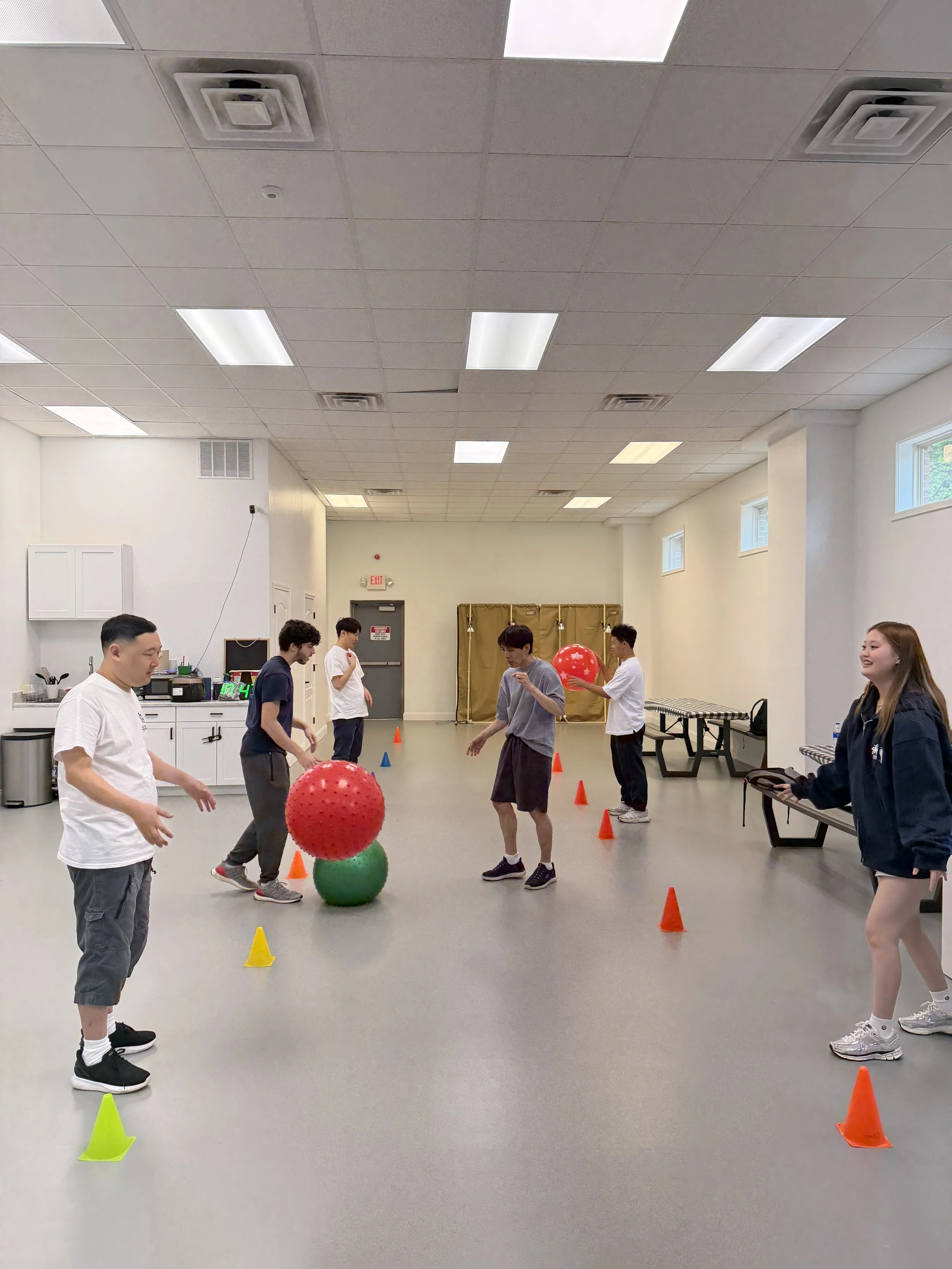 Group of young people participating in a game in an indoor gym or recreation room, with cones and large red and green balls, some people smiling and observing.