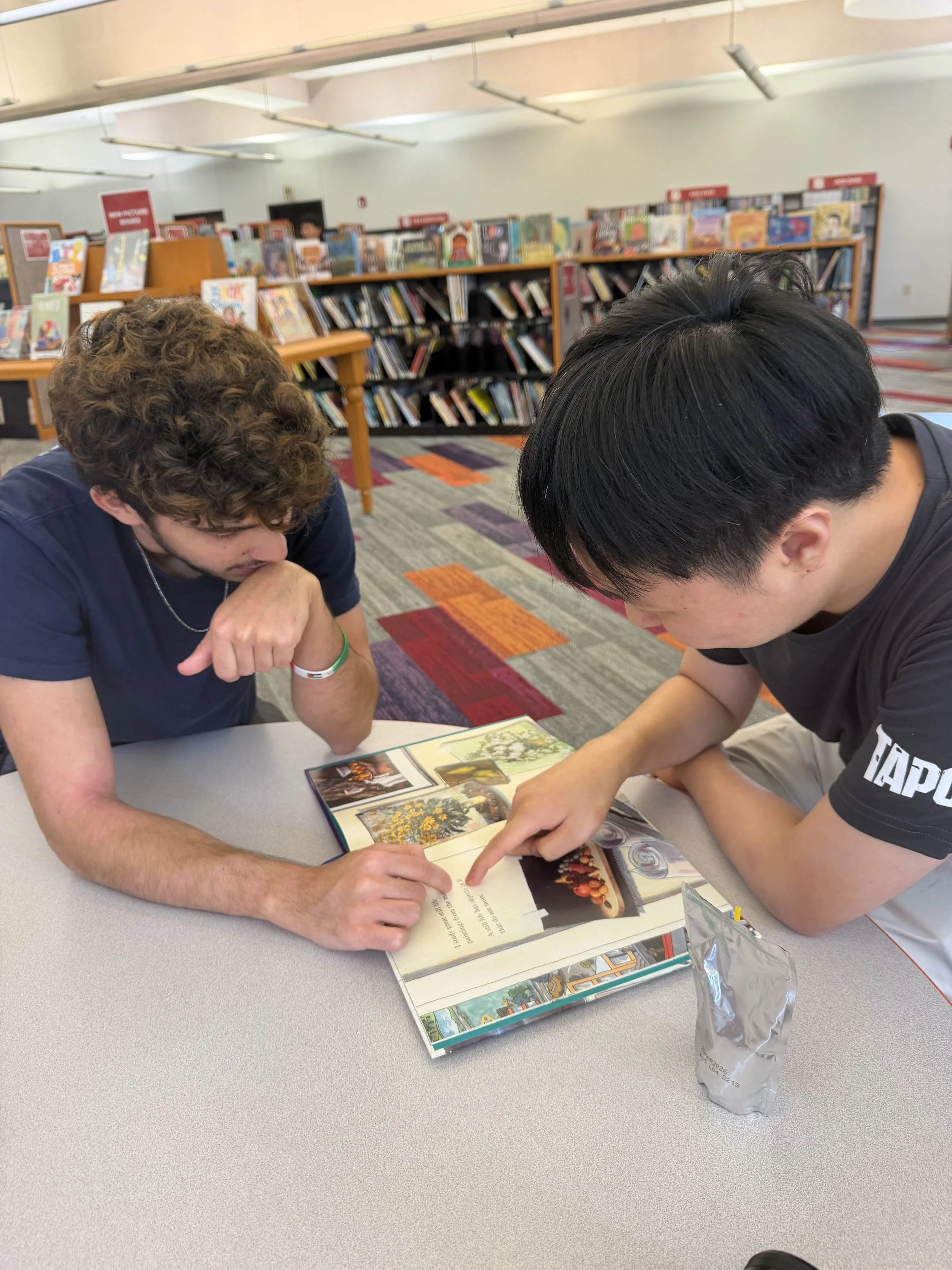 Two young men sitting at a table in a bookstore, looking at a book together. The bookstore has shelves filled with books in the background, and a bag of snacks is on the table.