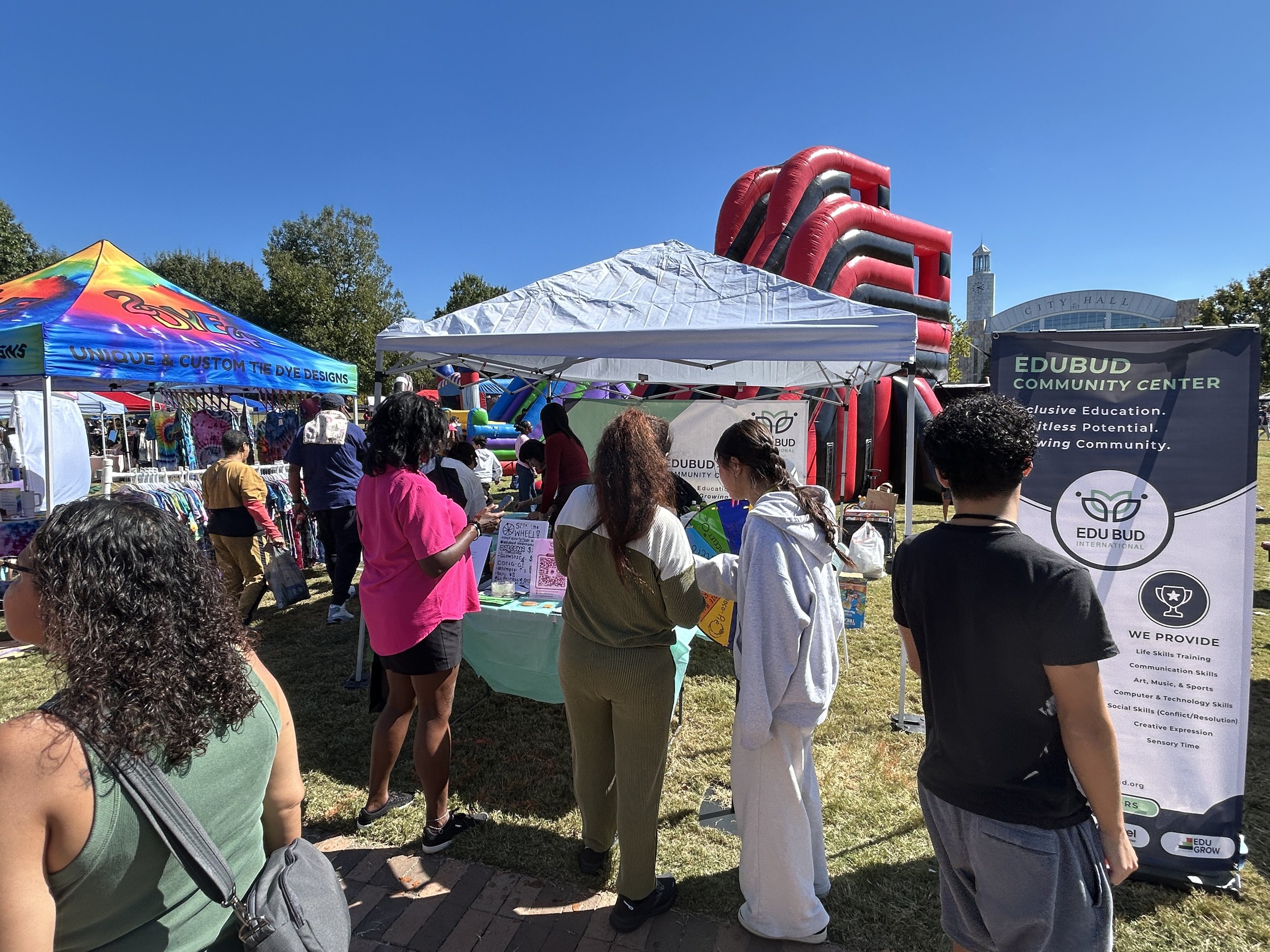 Crowd of people attending an outdoor community event around white tents and booths, with a large inflatable slide in the background and a city hall building visible against a clear blue sky.