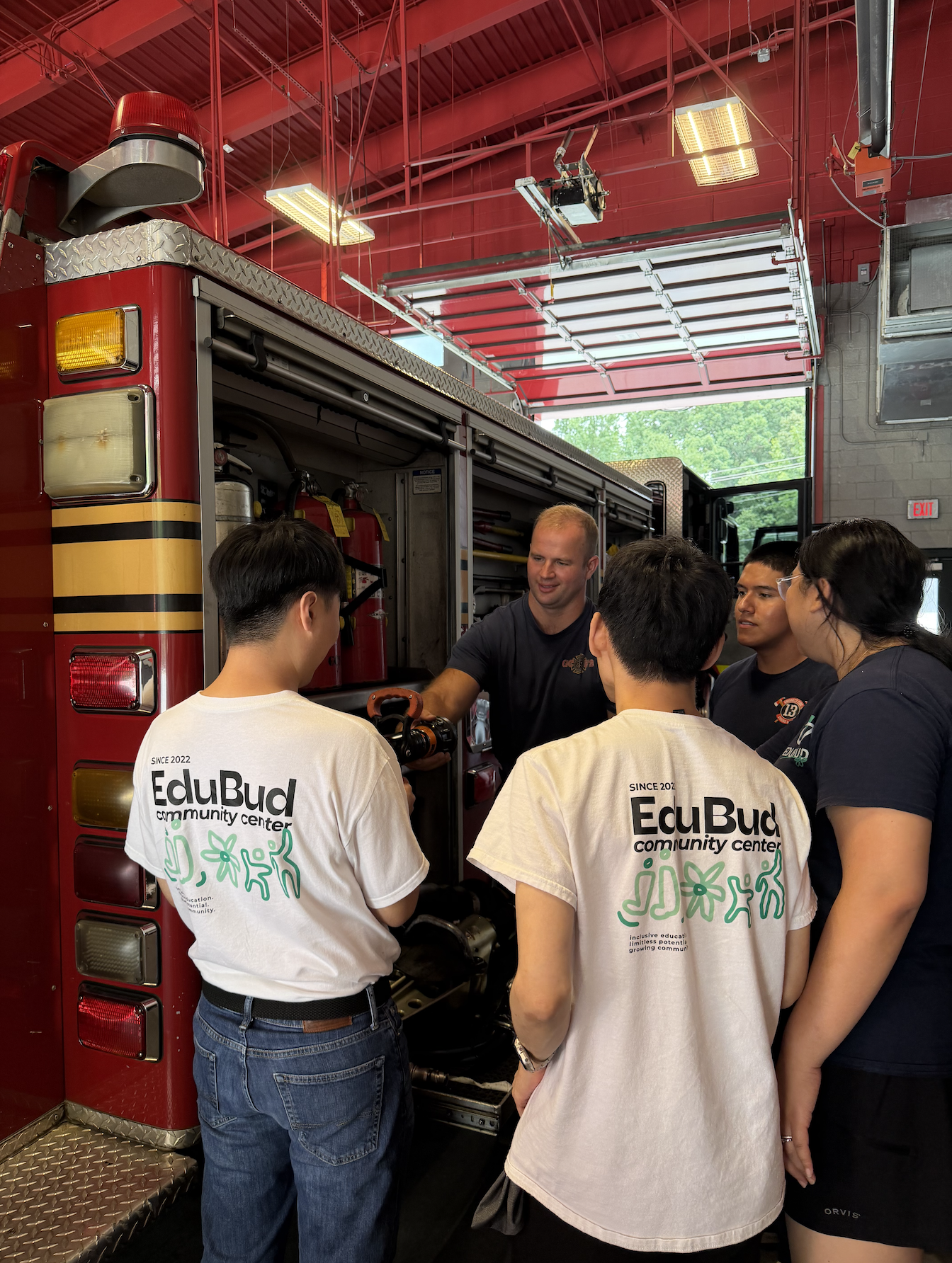 A group of students wearing EduBud community center T-shirts gathered around a firefighter inside a fire station, listening to the firefighter explain fire equipment from a fire truck.