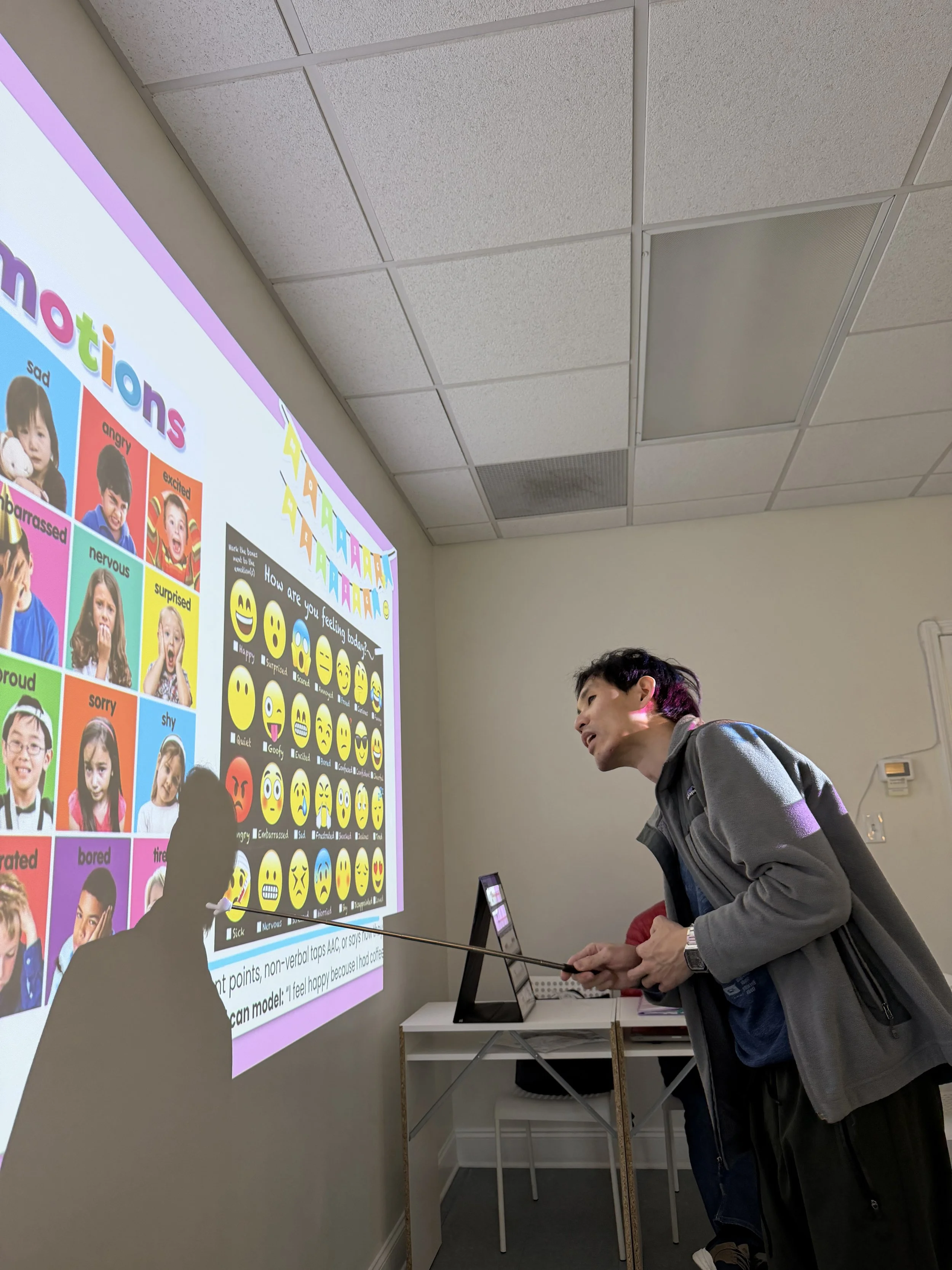 A person standing in front of a presentation slide with colorful emotion images and words on a classroom or lecture room wall.