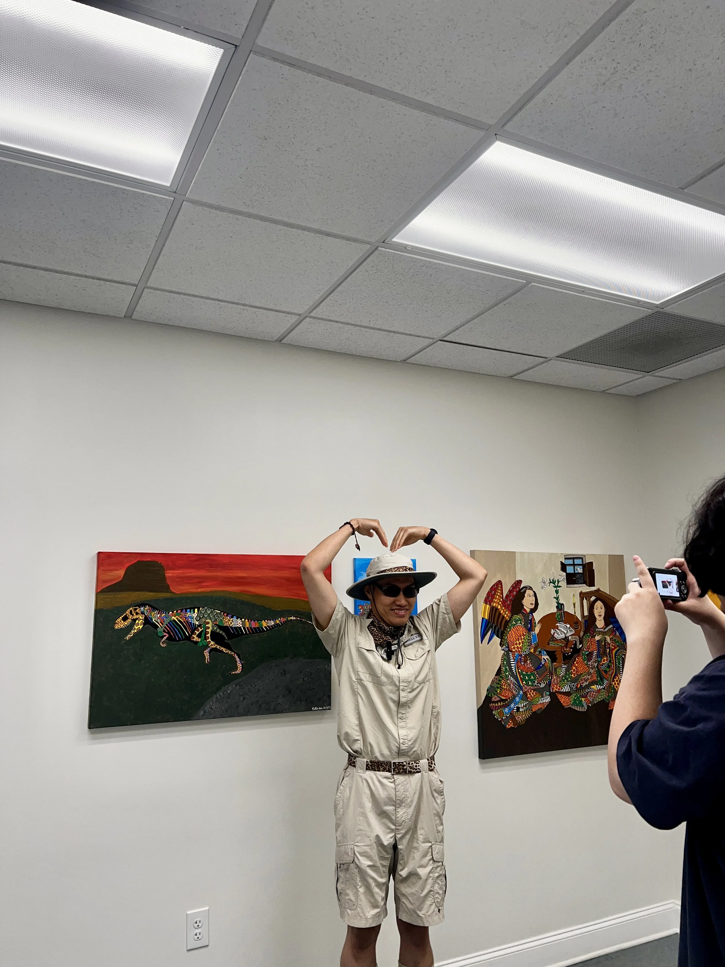 A person standing in front of colorful artwork and making a heart shape with their hands, while a person on the right takes a photo.