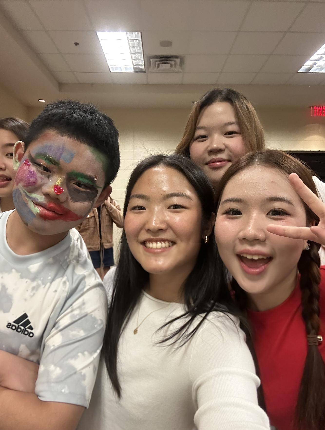 Group of young people taking a selfie indoors, smiling, with one person wearing colorful face paint resembling a clown or joker.
