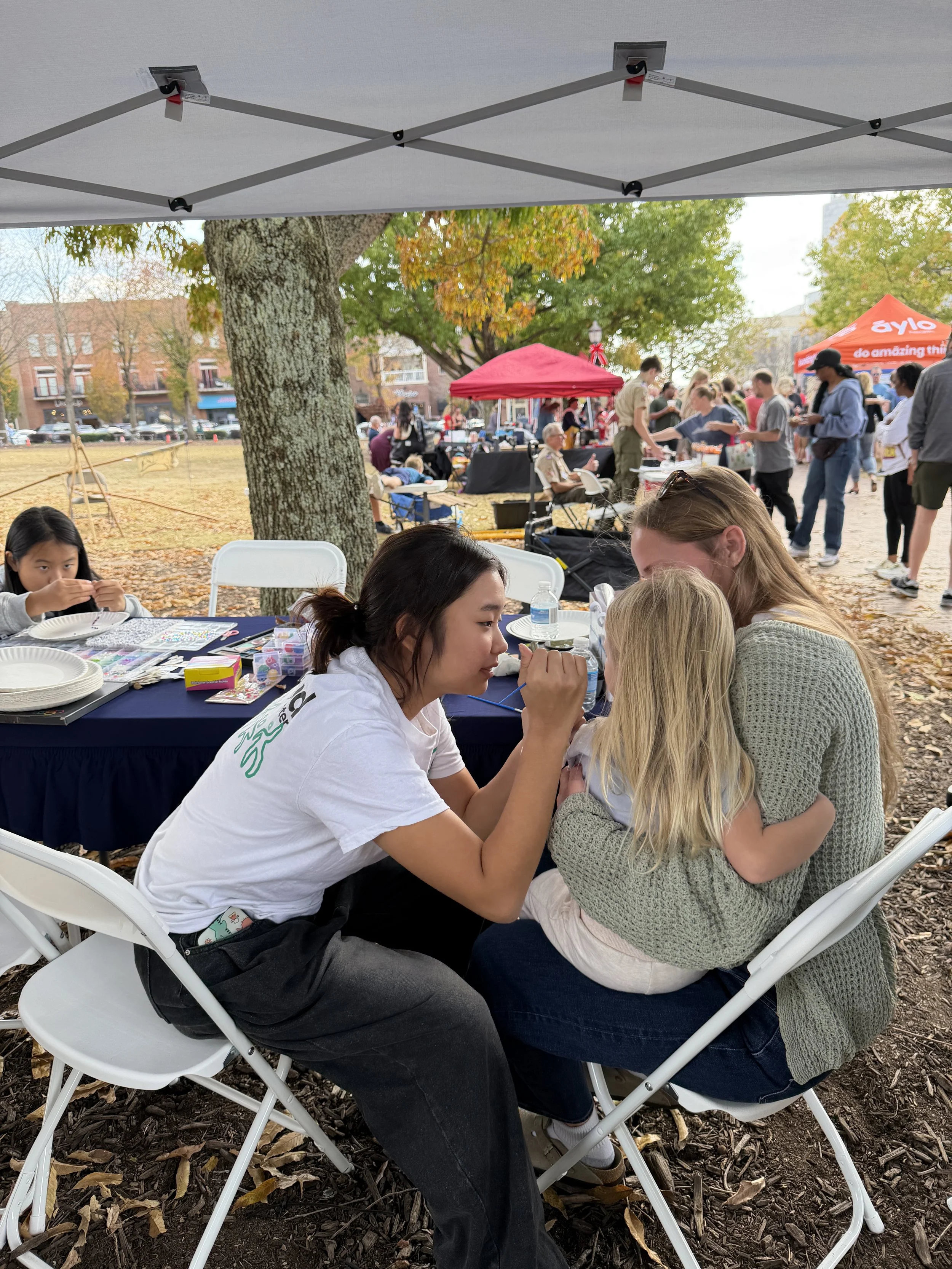 A woman with dark hair and a white shirt is drawing on a young girl's face, while another woman with blonde hair holds the girl. They are sitting at a table under a tent at an outdoor event during autumn. The background shows a park with trees, other