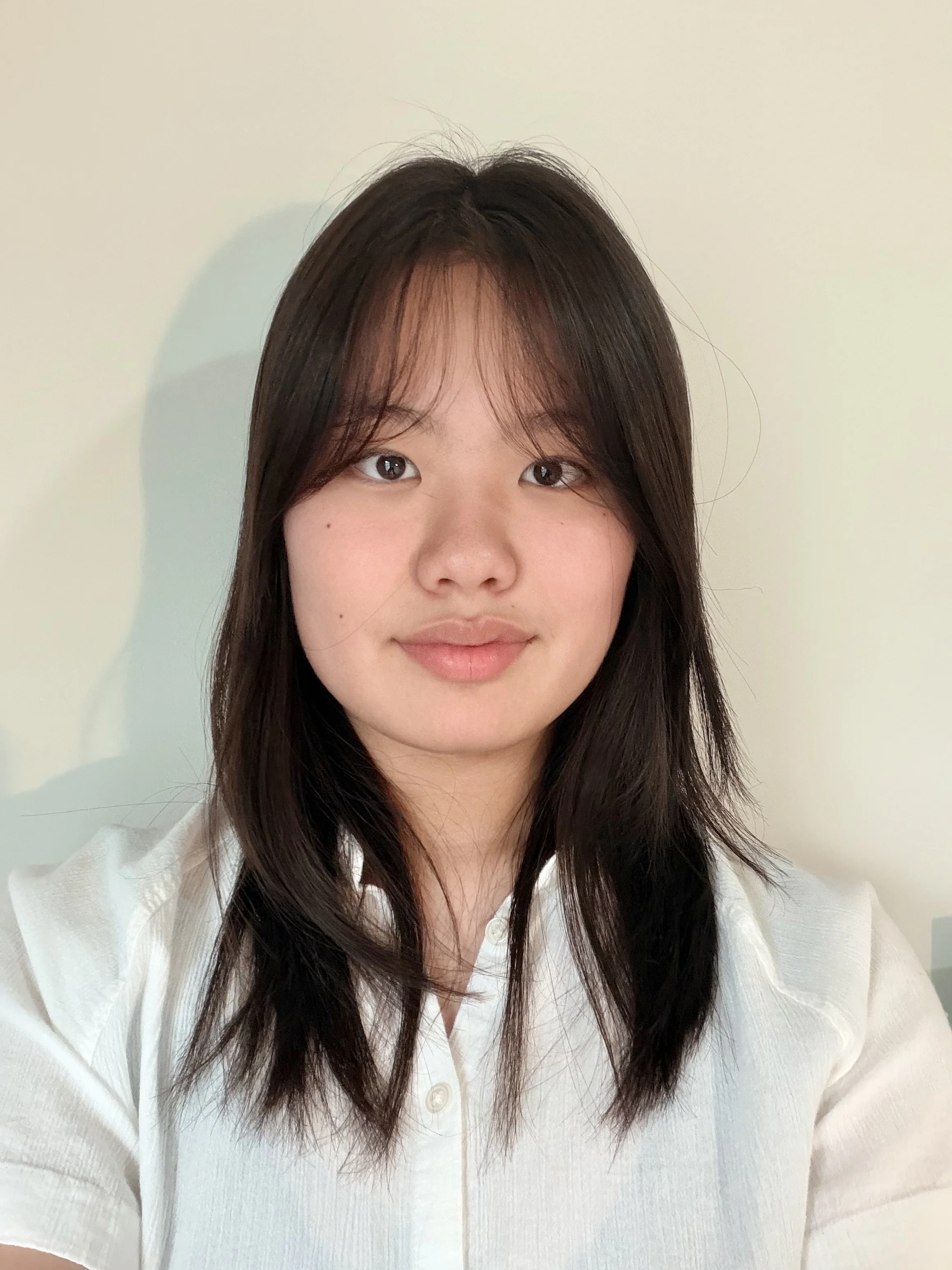 A young woman with long dark hair wearing a white shirt, taking a selfie against a plain off-white wall.