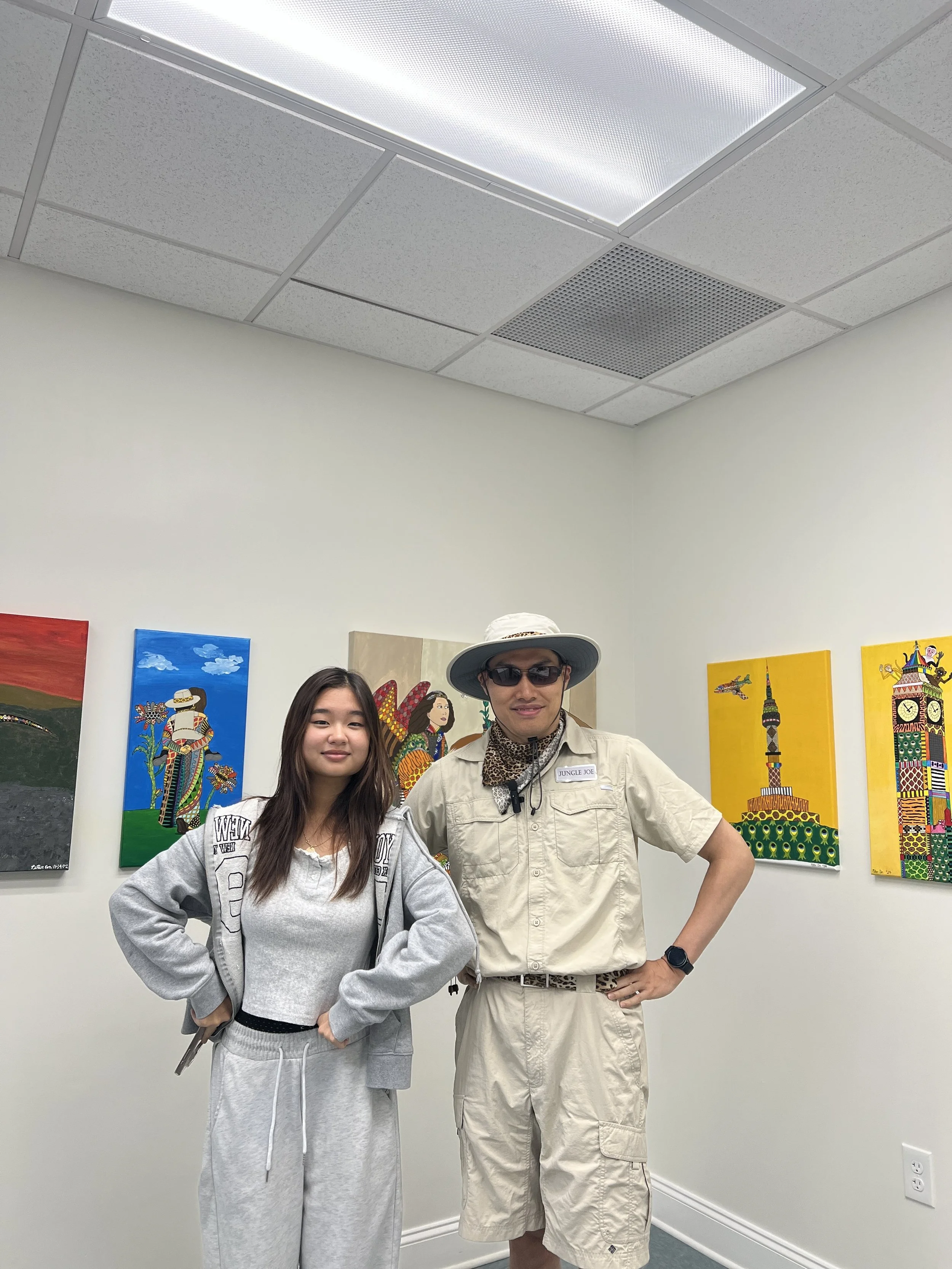 Two people standing in front of colorful artwork at an art gallery. The person on the left is a young woman with long dark hair, wearing a gray hoodie and sweatpants. The person on the right is an older man wearing sunglasses, a wide-brimmed hat, a b