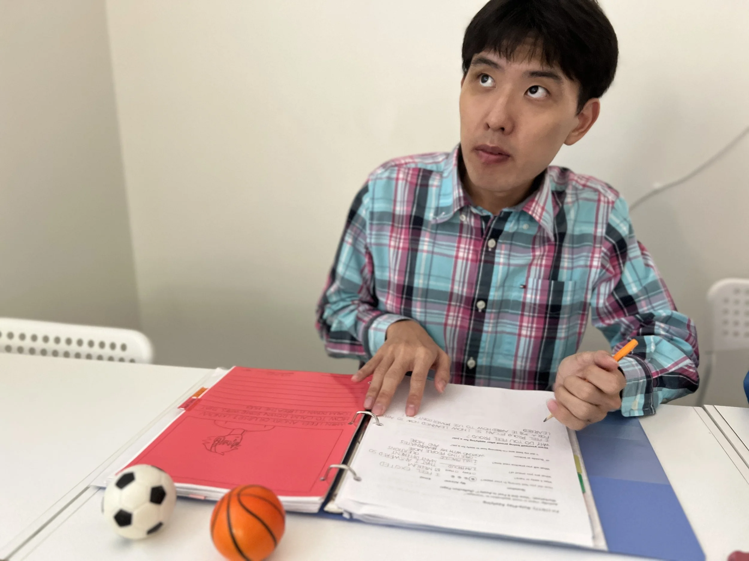 A young man with dark hair wearing a colorful plaid shirt sitting at a white table with a red binder, papers, and writing utensils. In the foreground are small soccer balls in black and white, orange, and black. The background is a simple off-white w