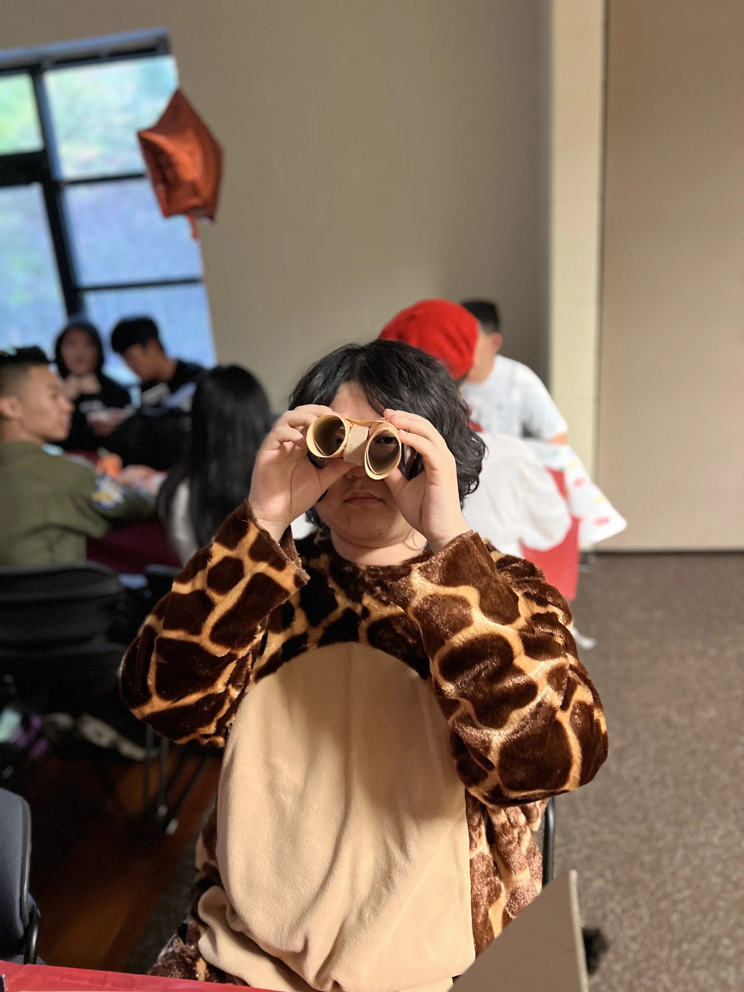 A child wearing a giraffe costume holding binoculars up to their eyes in a room with other people in the background.