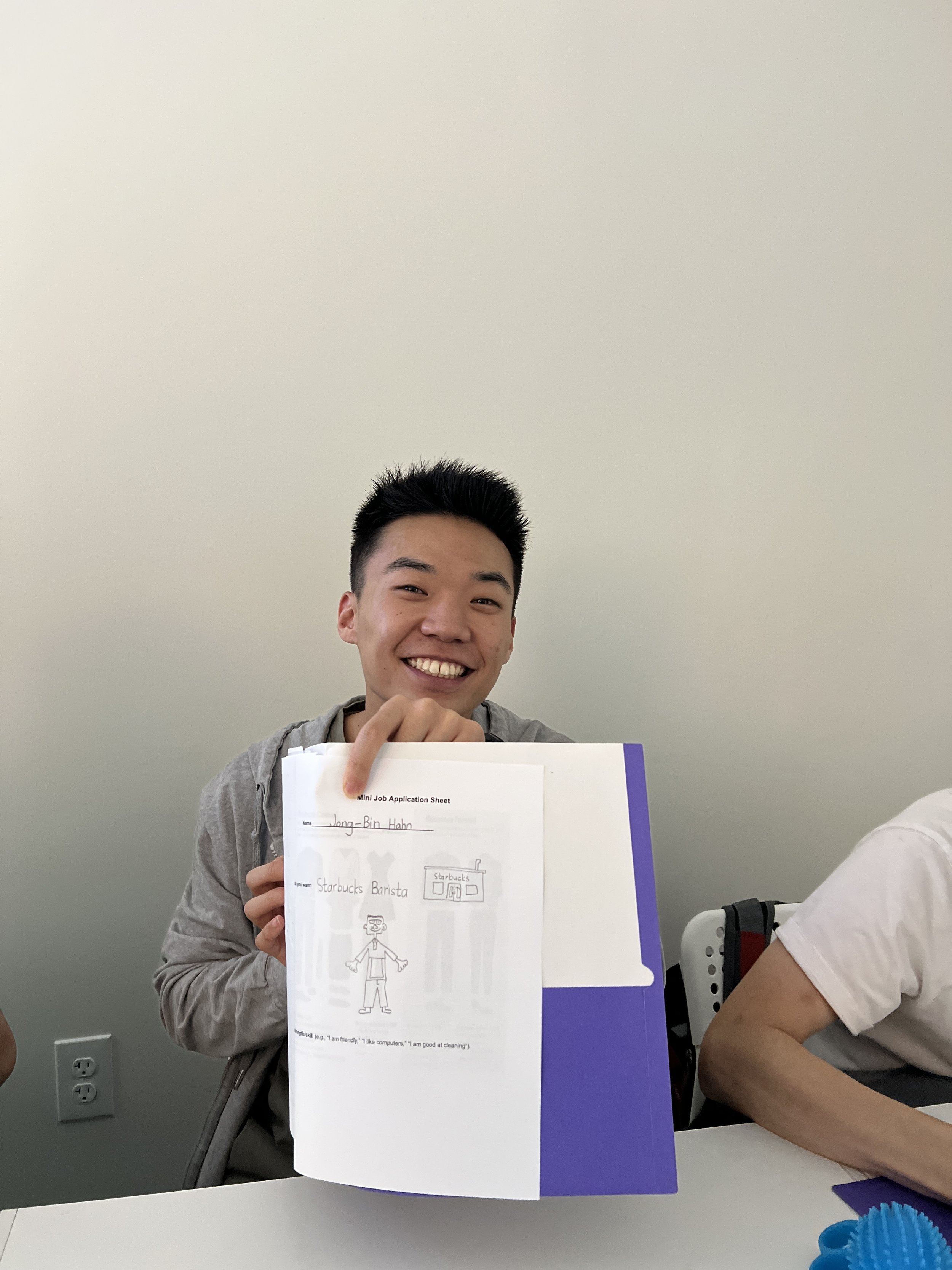 A young man smiling and holding a sheet of paper with a drawing of himself, in a classroom or office setting.