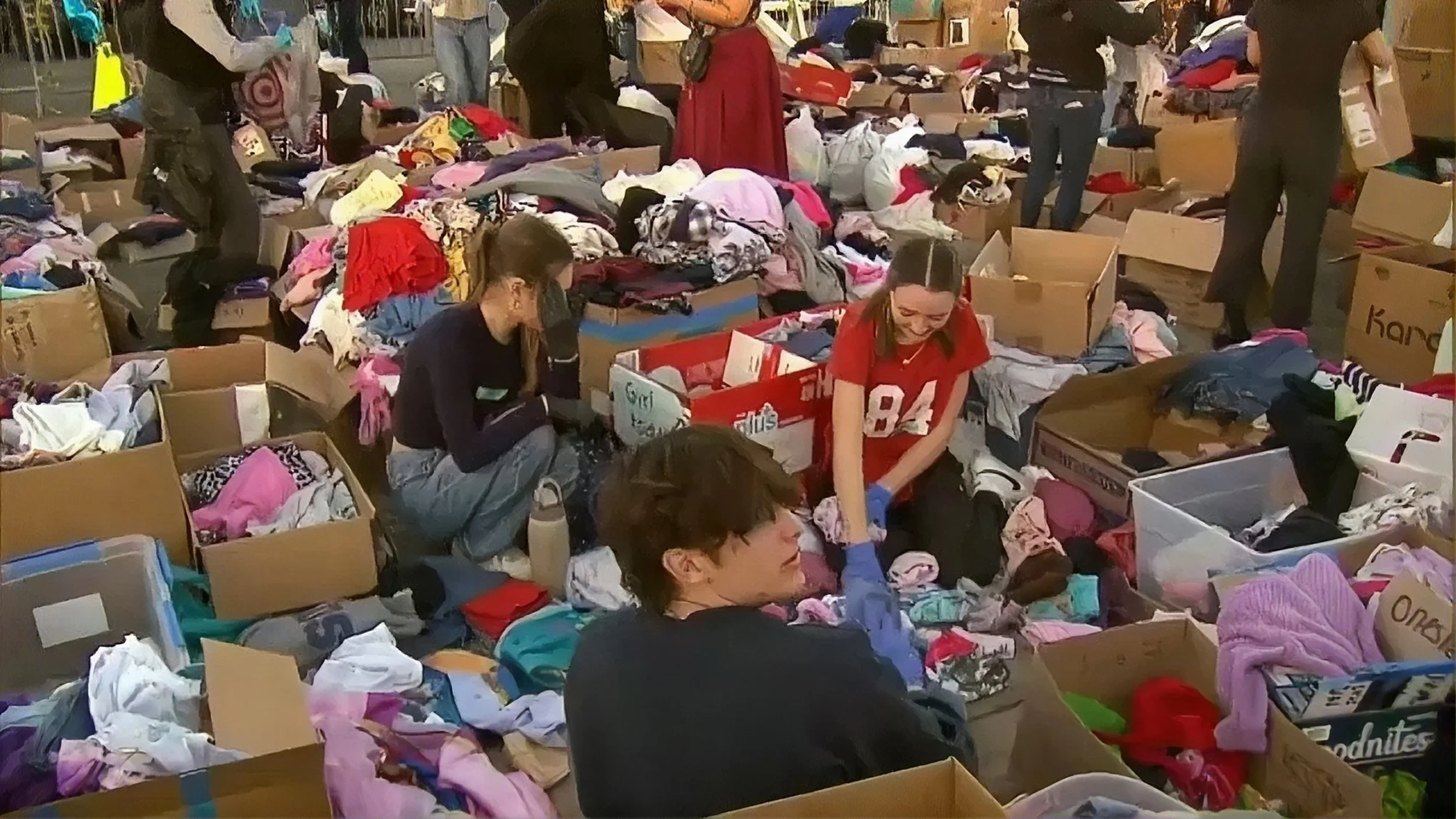 People sorting and organizing clothes in a large indoor space, surrounded by numerous boxes and piles of clothing.