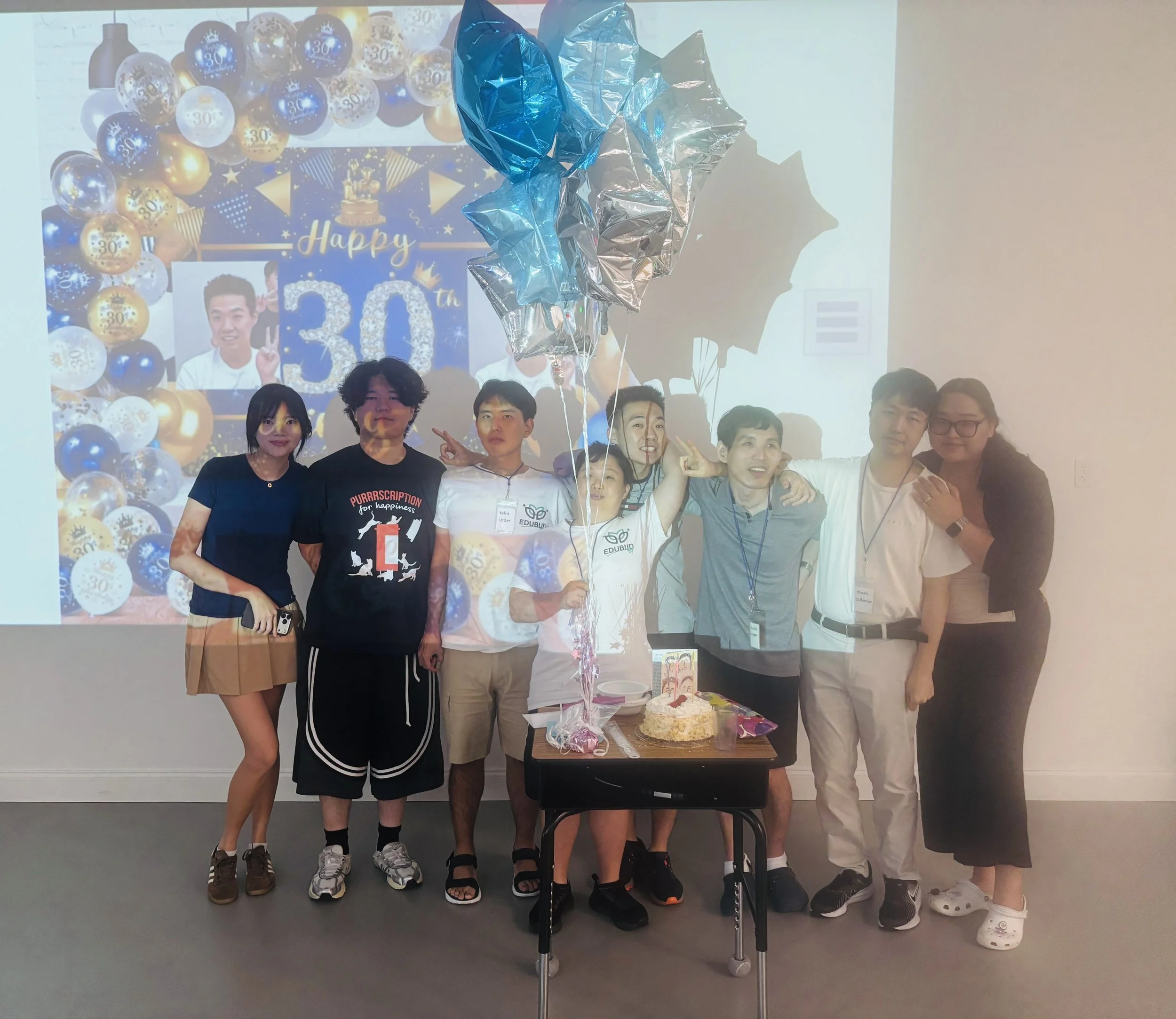 A group of people celebrating a birthday or special occasion, standing behind a table with a cake and party decorations, including balloons and a birthday banner in the background.