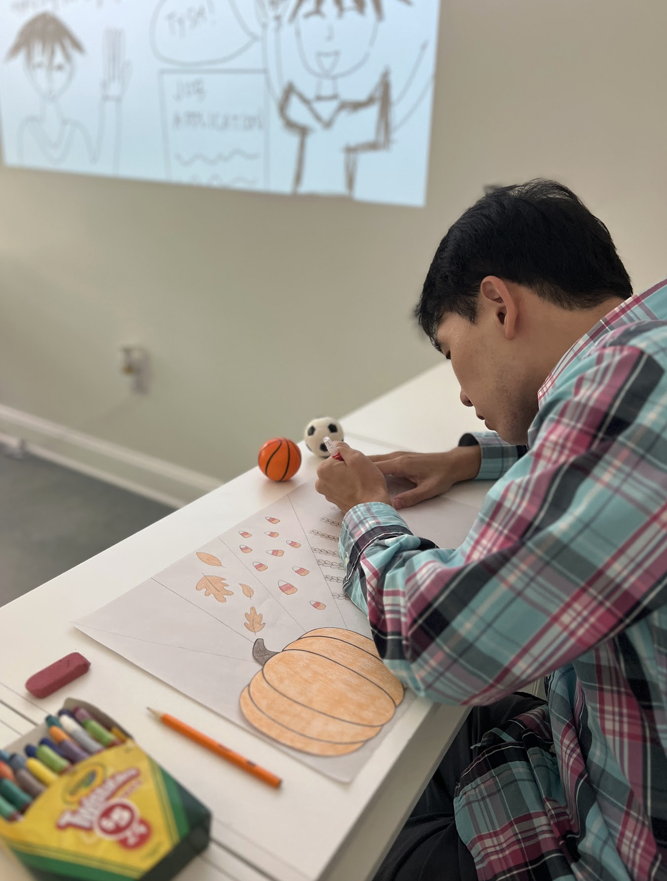 A person seated at a white table coloring pumpkin and leaf drawings with orange crayon, at a classroom or art room. Art supplies and candies are on the table, with a background projection of a cartoon drawing of a person with short hair, an open mouth, and a speech bubble.