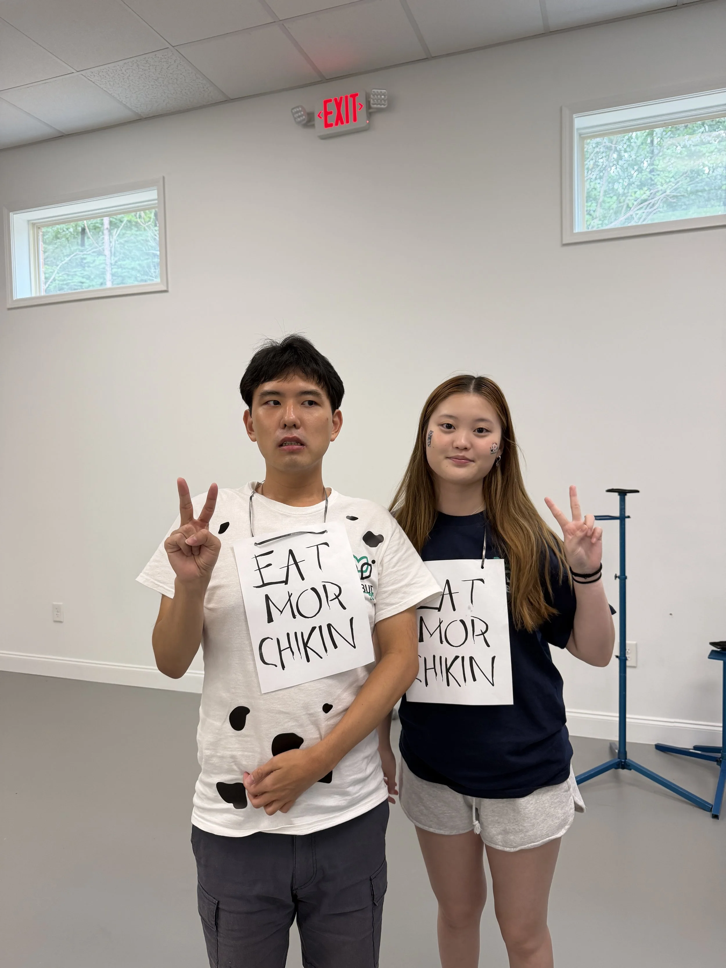 Two young people standing indoors with white walls and small rectangular windows, holding signs that say 'EAT MOAR CHIKIN' and making peace signs.