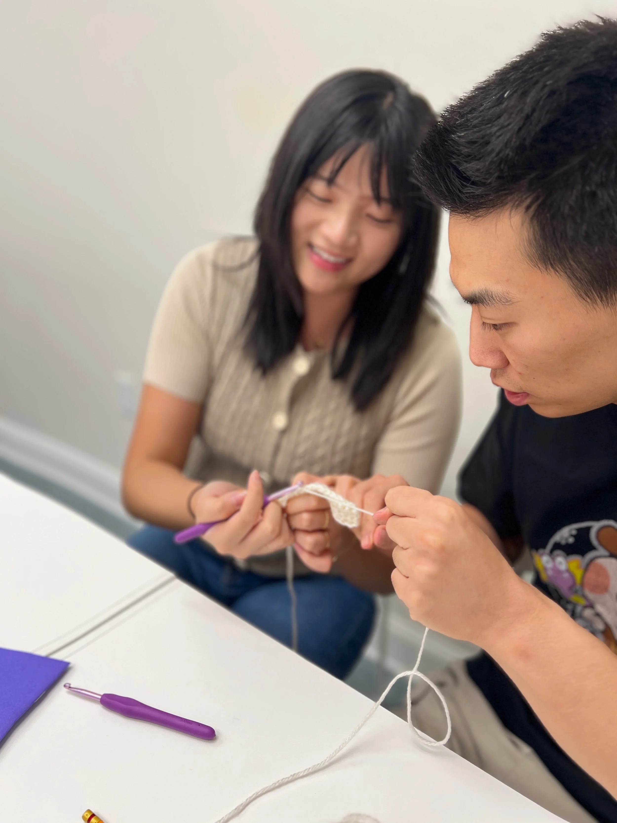 Two people knitting together at a table, one person with short dark hair and a woman with long dark hair smiling, with knitting supplies on the table.
