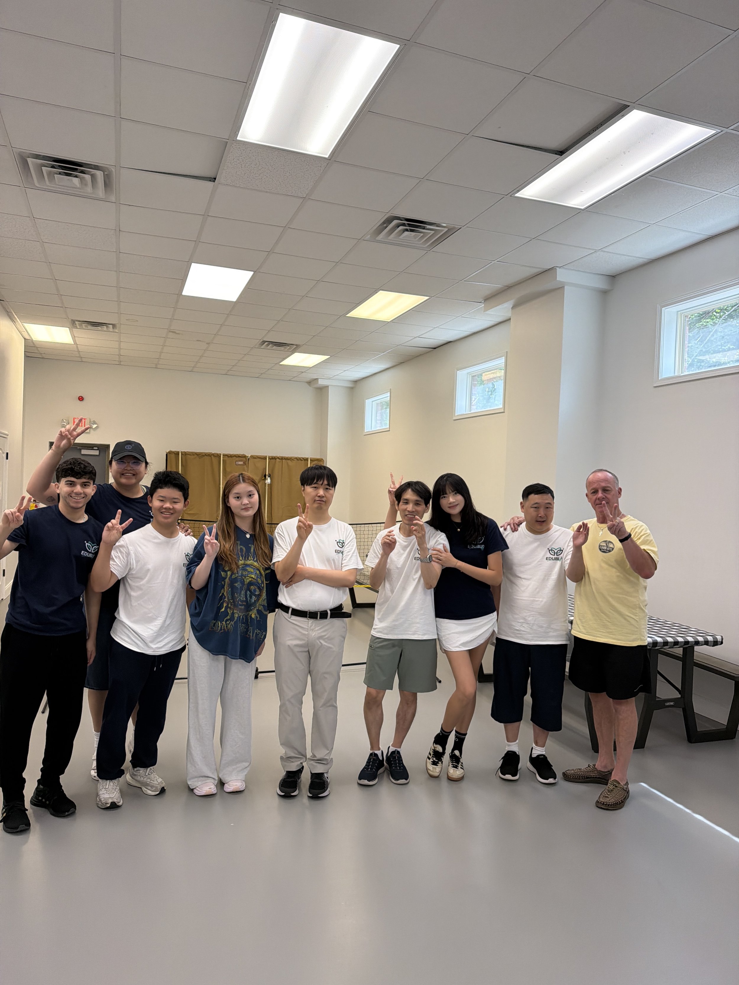 Group of young students and an older man posing together in a room with white walls and windows, some making peace signs.