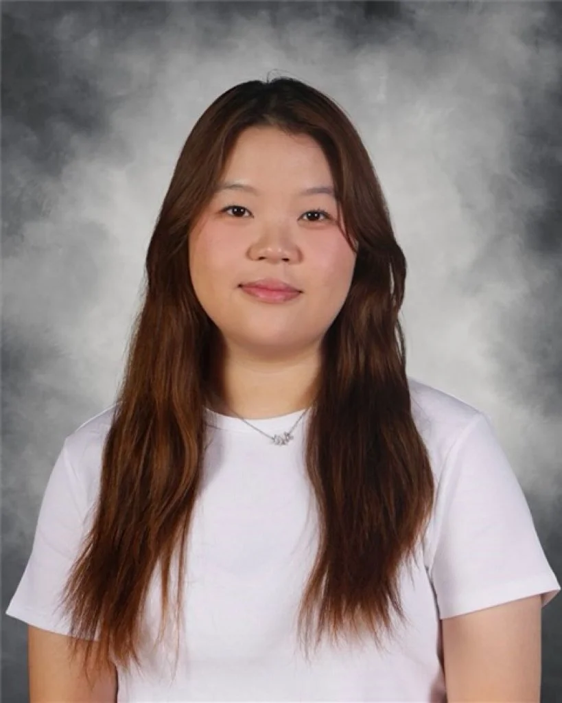 A young woman with long brown hair, wearing a white t-shirt and a necklace, smiling slightly against a gray textured background.