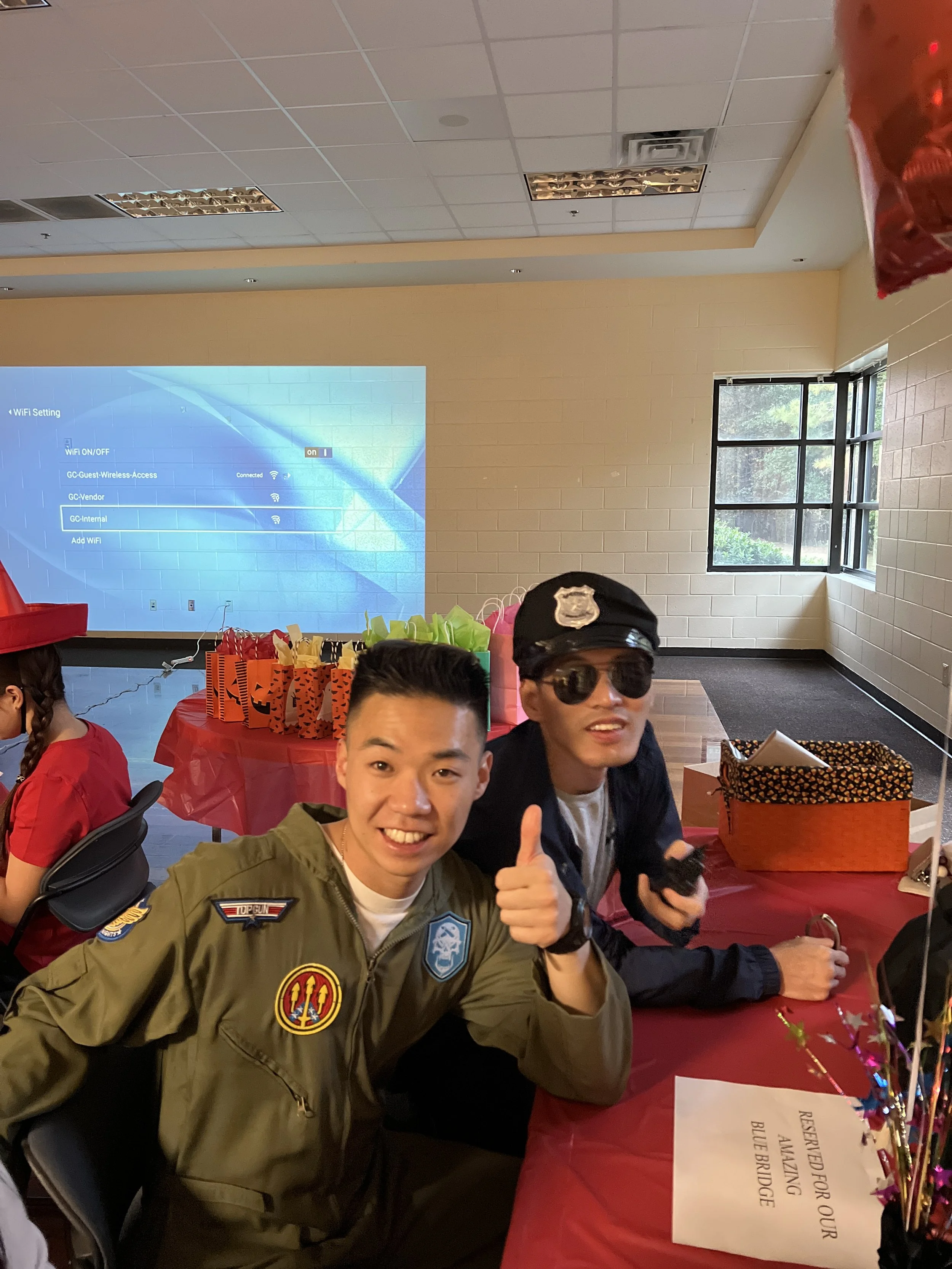 Two young men sitting at a table decorated for a celebration, with gift bags and party decorations. One man is wearing an aviation-themed jacket, sunglasses, and giving a thumbs-up. The other is wearing a black jacket and holding a phone. There are t