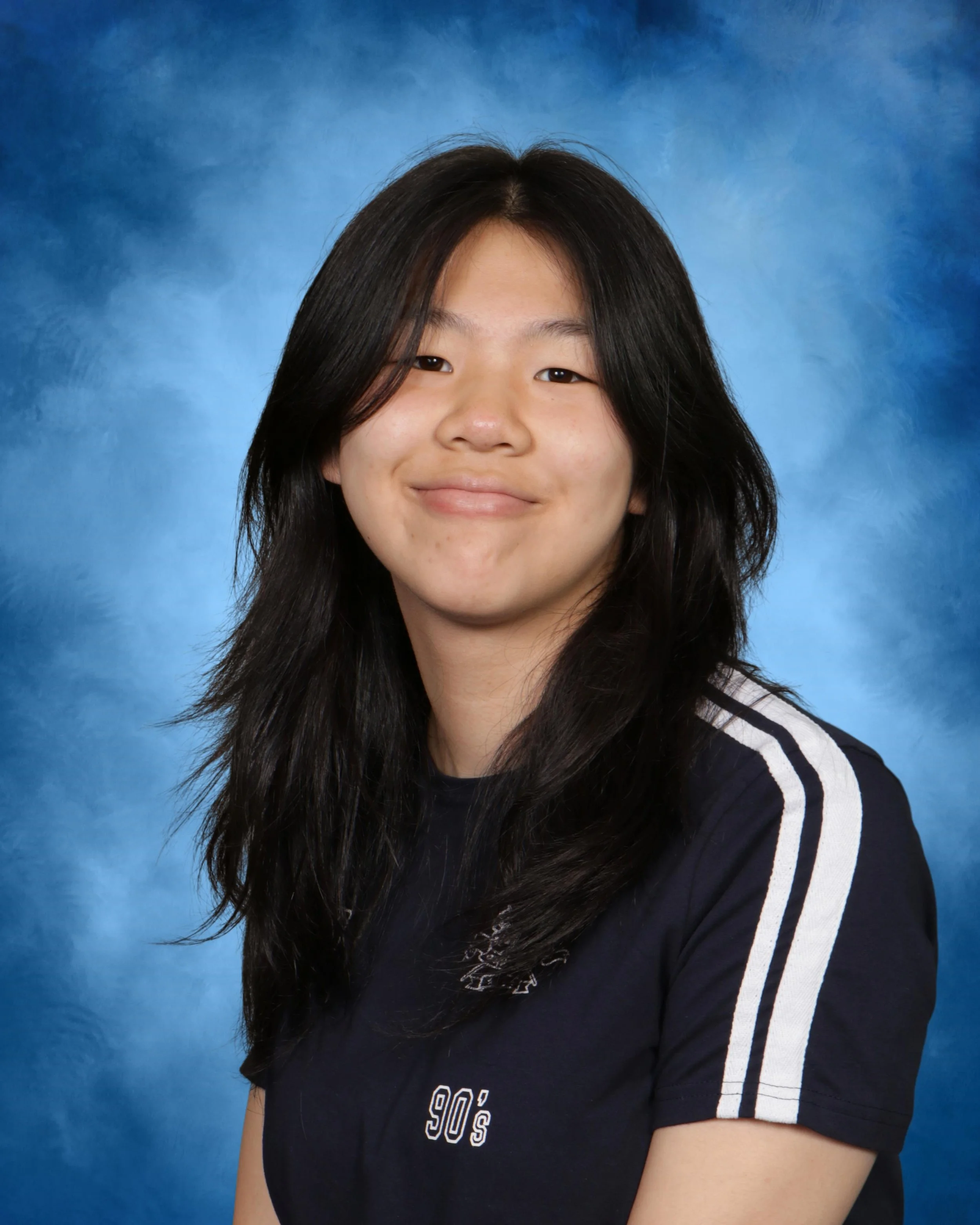 Close-up of a young person with long black hair, smiling, wearing a black T-shirt with white text and graphics, against a blue textured background.