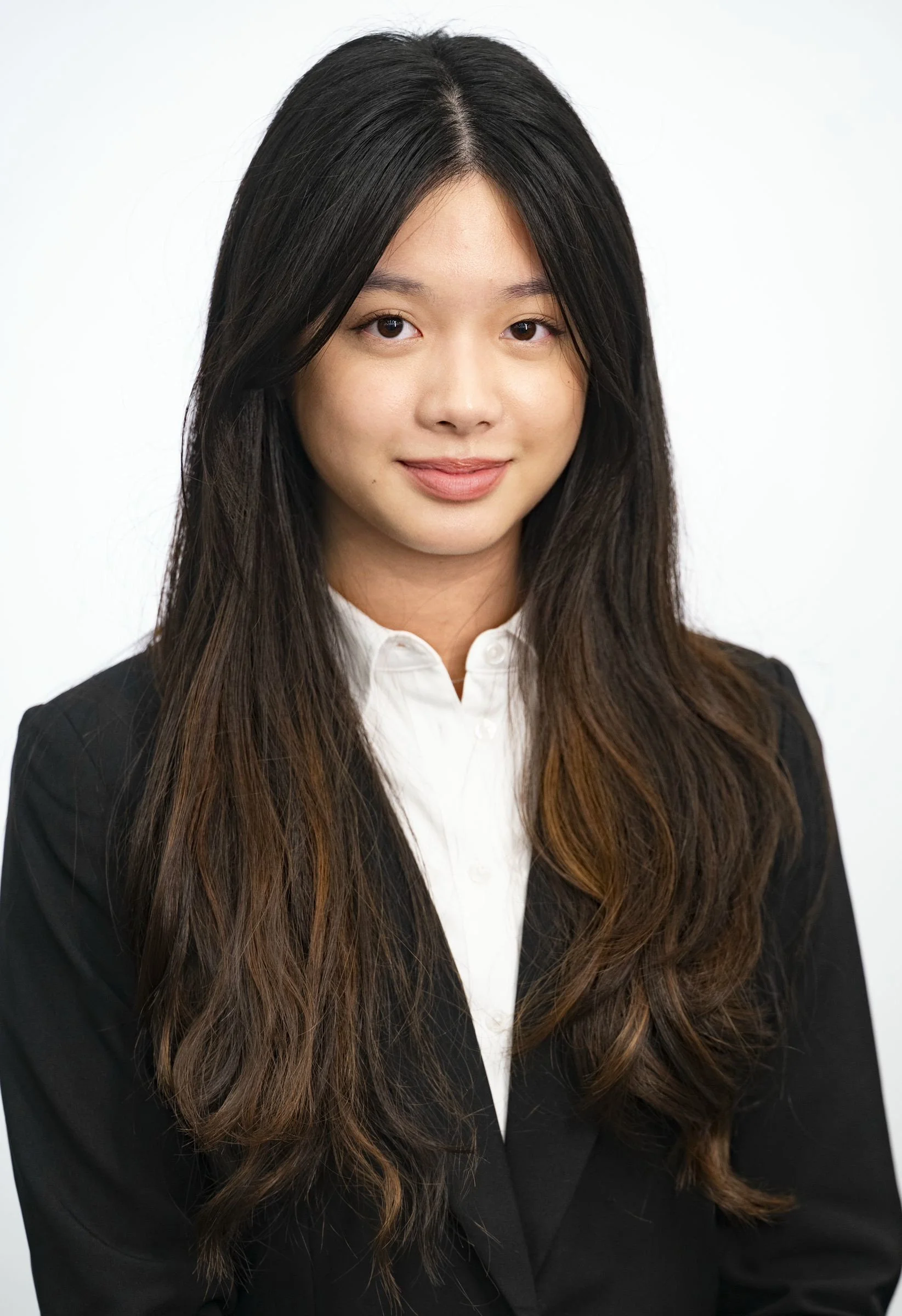 A professional woman with long dark hair, wearing a black blazer and white shirt, posing against a plain light background.