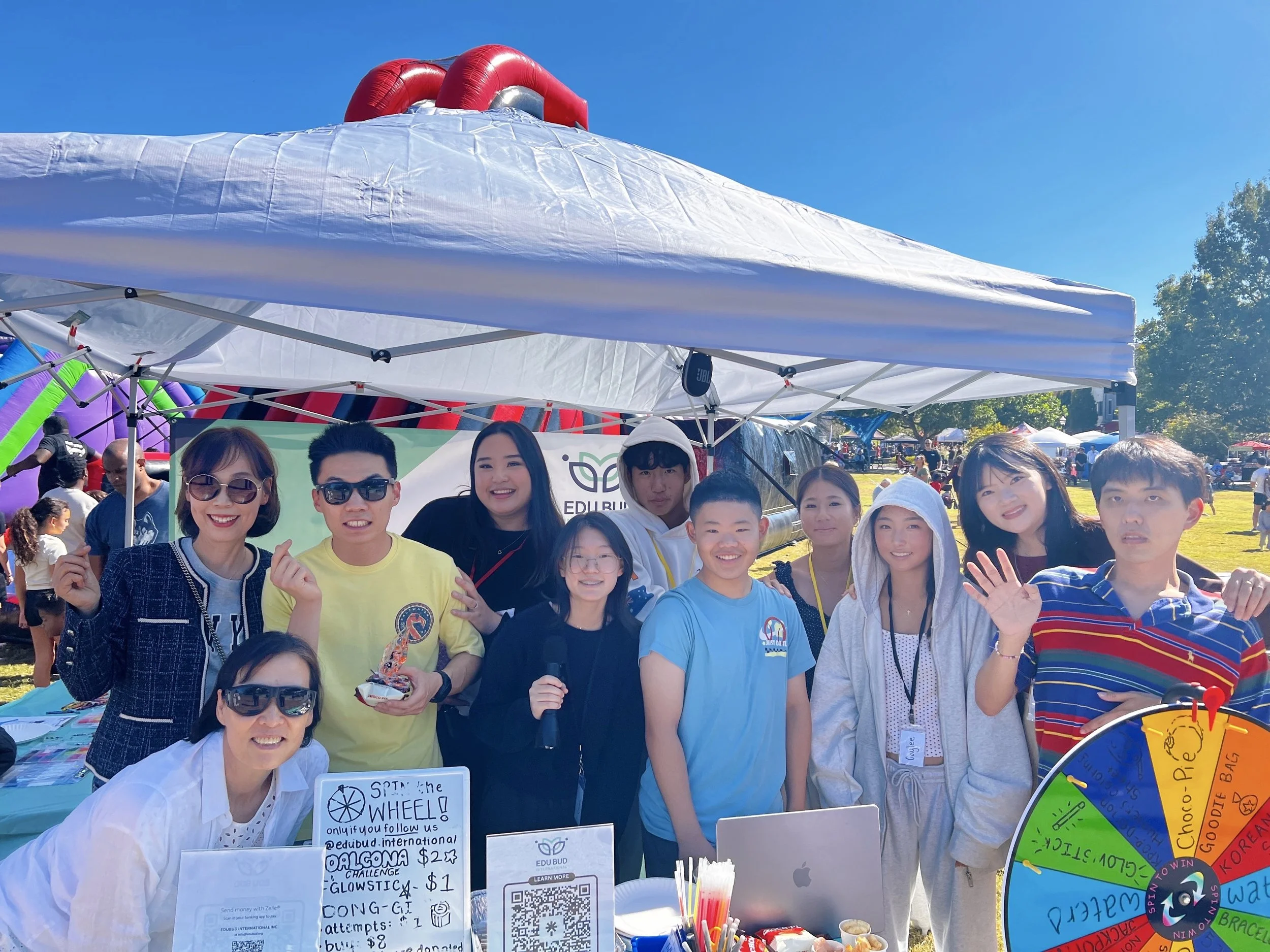 A group of people standing under a white canopy at an outdoor event with a clear blue sky in the background. Some people are smiling and looking at the camera, and there are various colorful signs and items on the table in front of them.