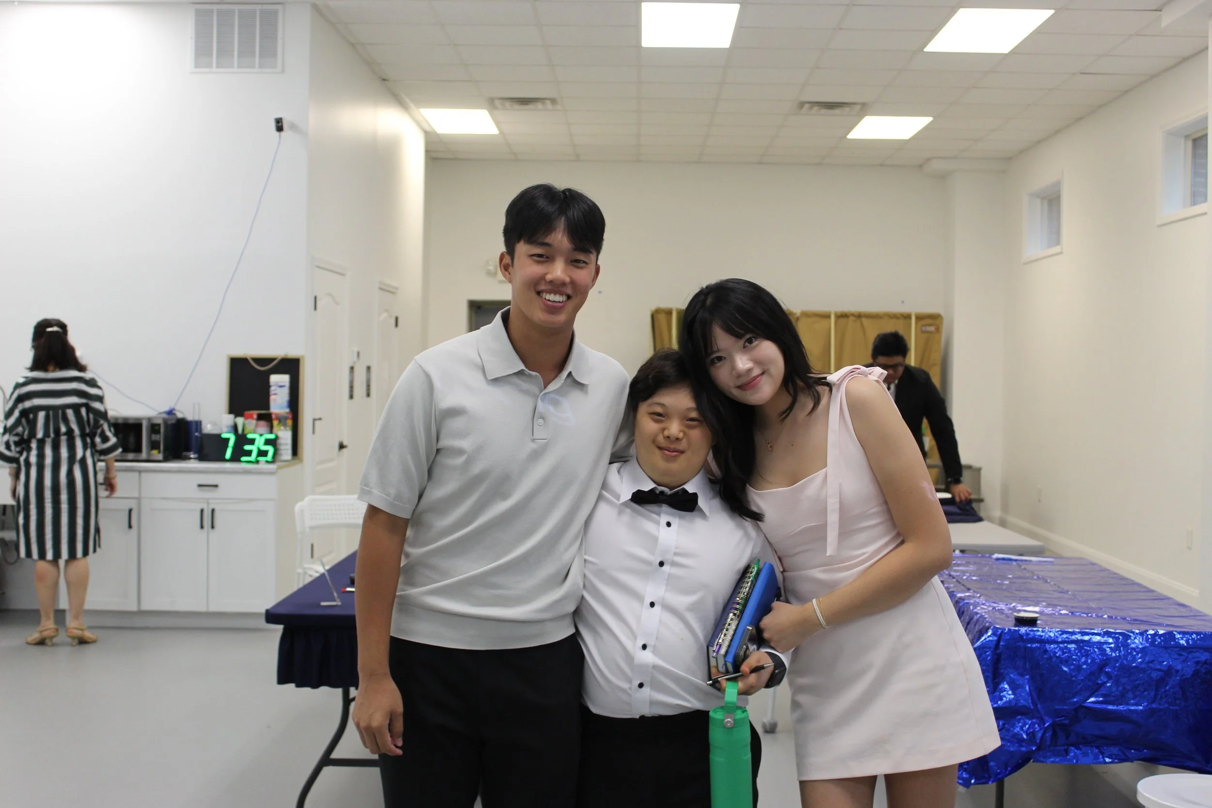 Four young people, two males and two females, are smiling and posing together in a well-lit room with white walls and a drop ceiling. They are dressed semi-formally, with the males in shirts and the females in dresses. One of the women has her arm ar