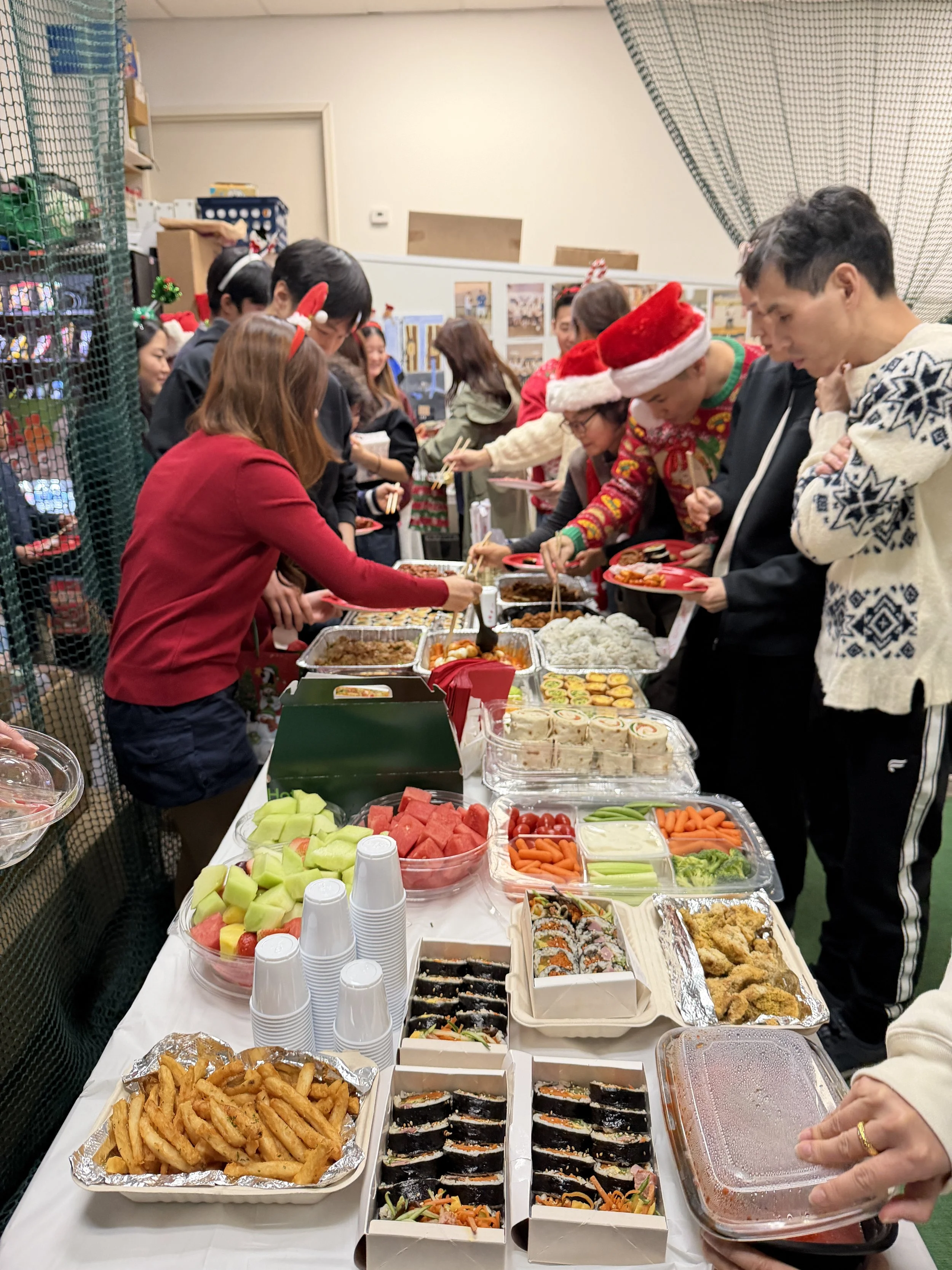People gathered around a table with various types of food, including sushi, fruit, vegetables, and fried snacks, during a holiday celebration with Christmas hats and decorations.