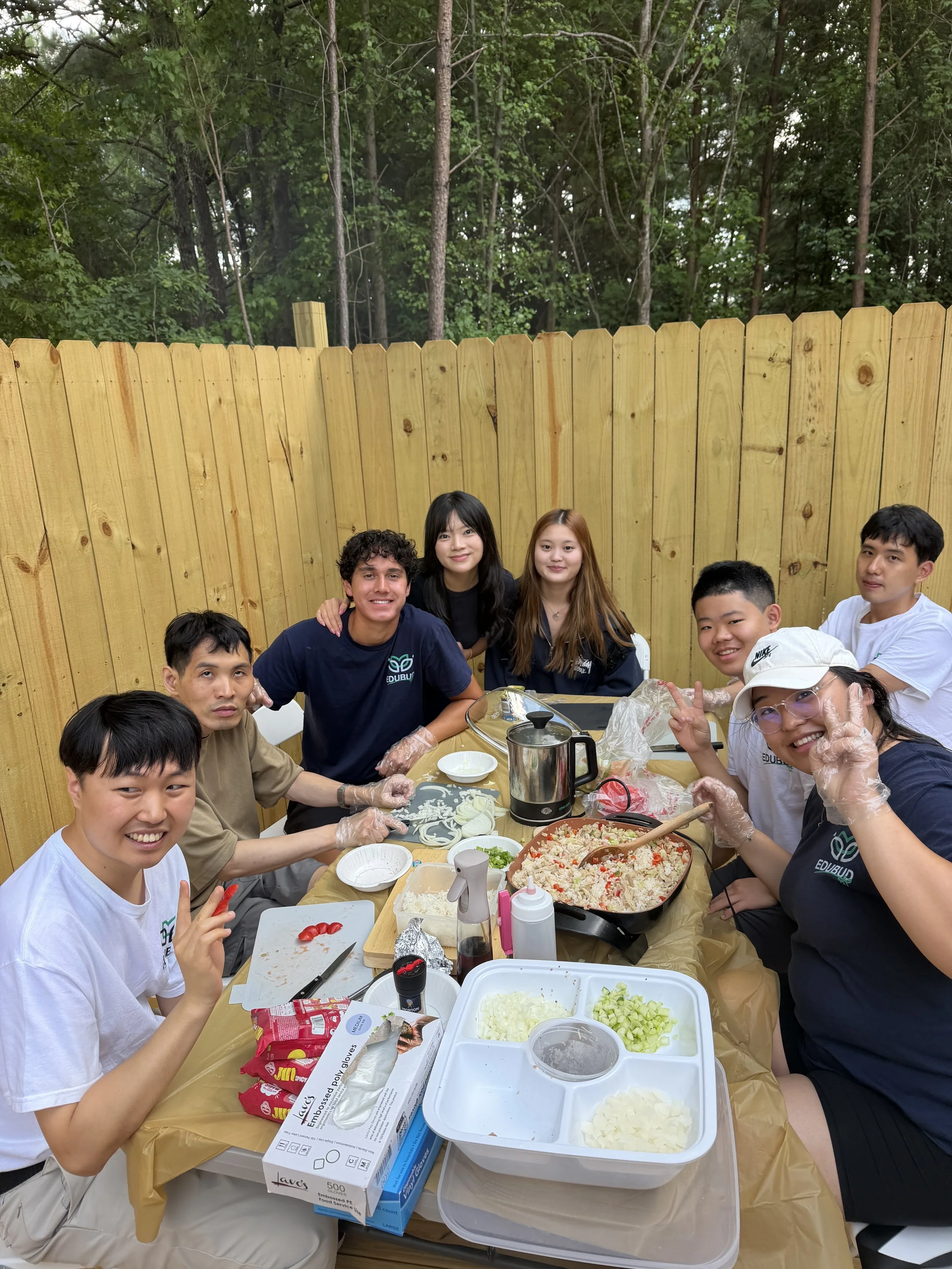 A group of people gathered around a table outdoors, enjoying a meal with various dishes, including chopped onions, diced cucumbers, and a large pot of rice salad. They are smiling, some making peace signs, and are wearing casual clothes and gloves. The background shows a wooden fence and trees.