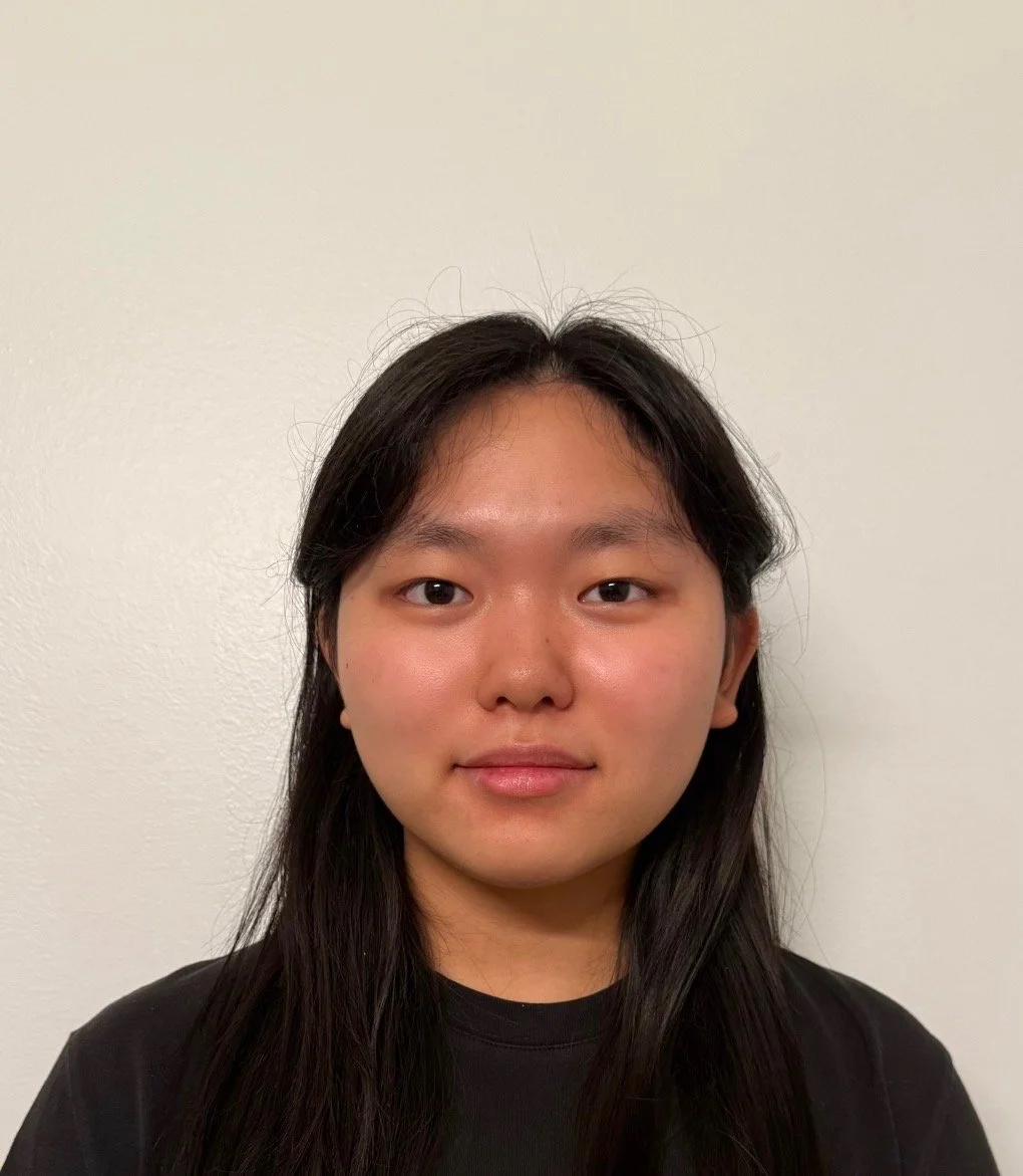 Front-facing portrait of a young woman with long black hair, wearing a black top, standing against a plain white wall.