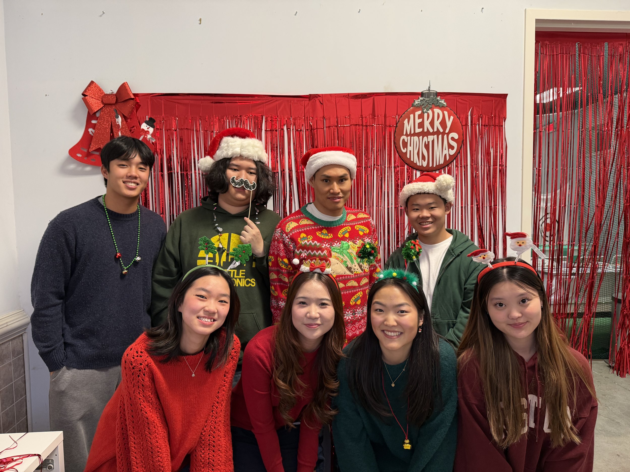 Group of young people celebrating Christmas, wearing Santa hats, festive sweaters, and holiday accessories, standing in front of a red Christmas backdrop with decorations.