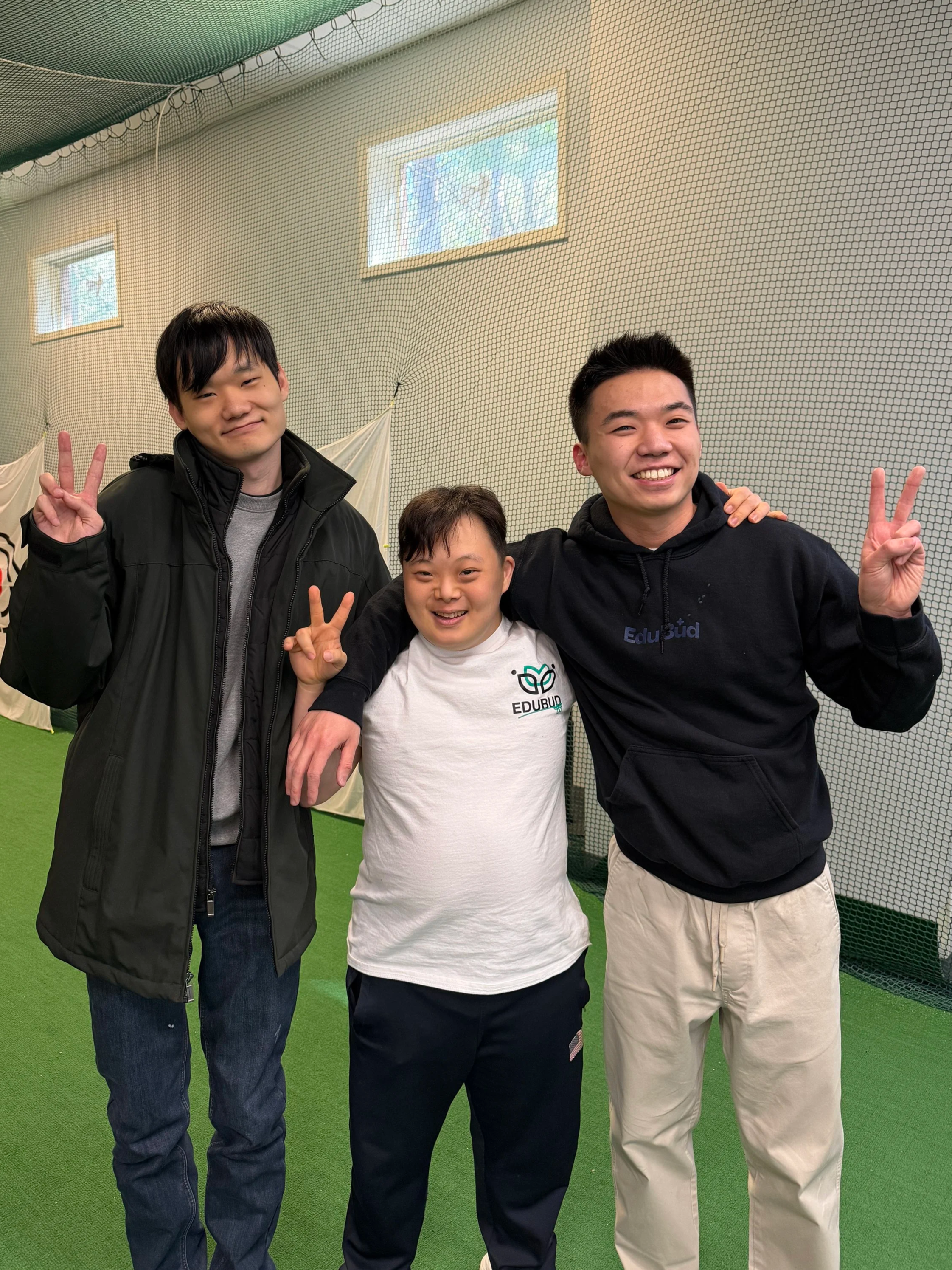 Three young men smiling and making peace signs with their fingers, posing in an indoor sports or recreation facility with green turf and a netted wall background.
