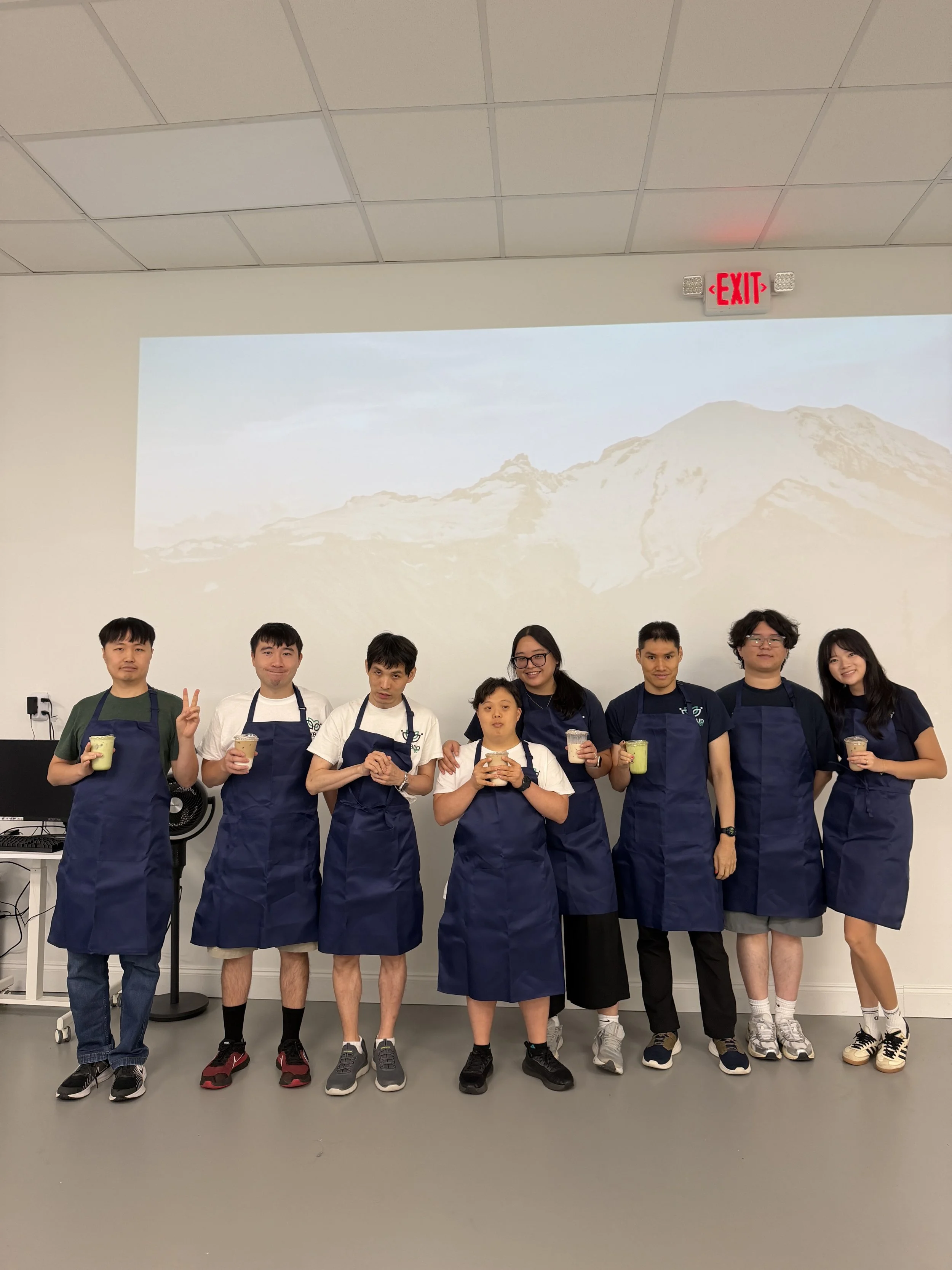 A group of nine people standing indoors, posing for a photo. They are wearing aprons and holding drinks, with a mountain landscape projected on the wall behind them.