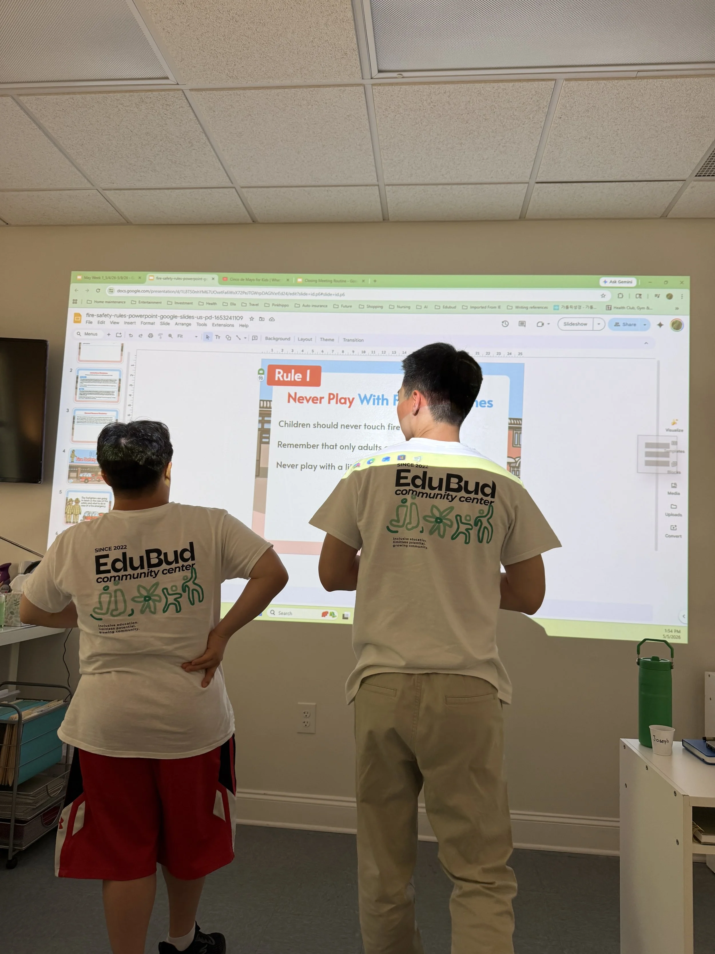 Two young men are standing in front of a large screen displaying a presentation slide titled 'Rule 1' about fire safety. The man on the right wears a white t-shirt with 'EduBud community center' printed on the back. The other person wears red shorts.