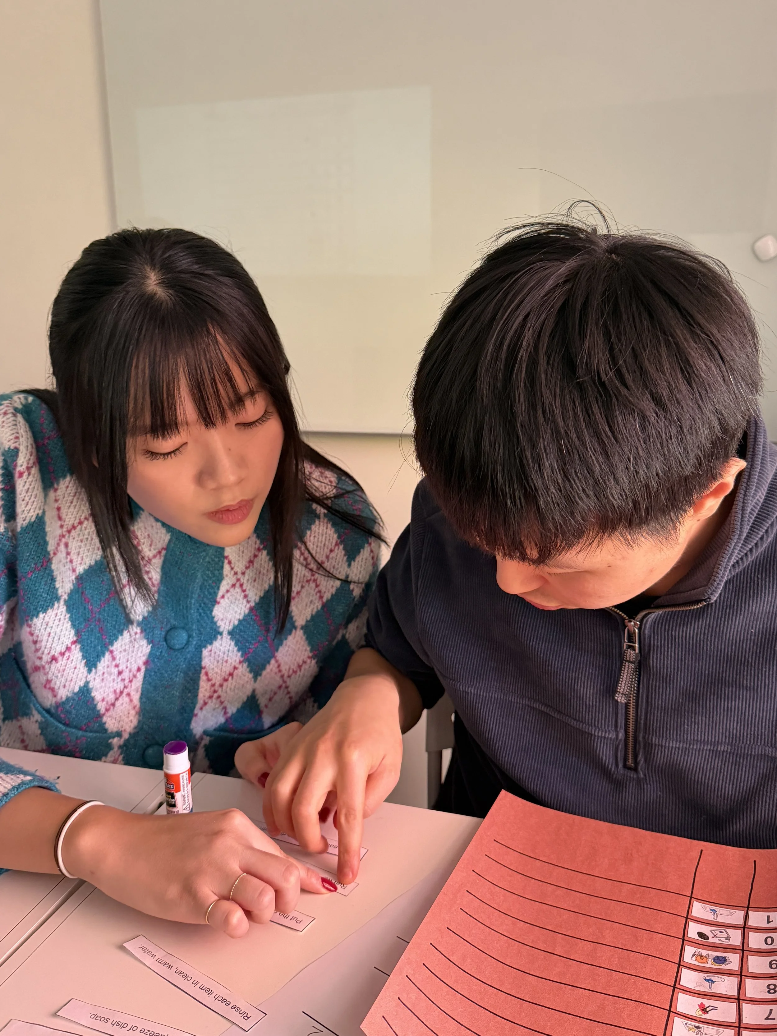 A young woman and a young man sit together at a table, engaged in a learning activity that involves pointing at laminated words and symbols on a sheet of paper and a colorful sheet with lines. The woman's hair is dark and straight, and she is wearing