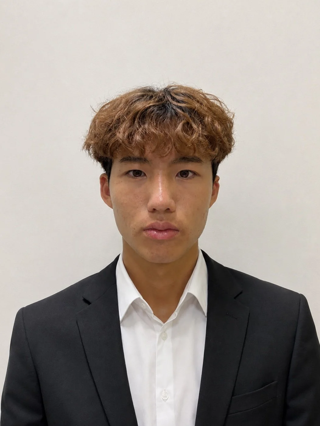 A young man with curly brown hair and light skin wearing a black suit jacket and white dress shirt standing against a plain white background.