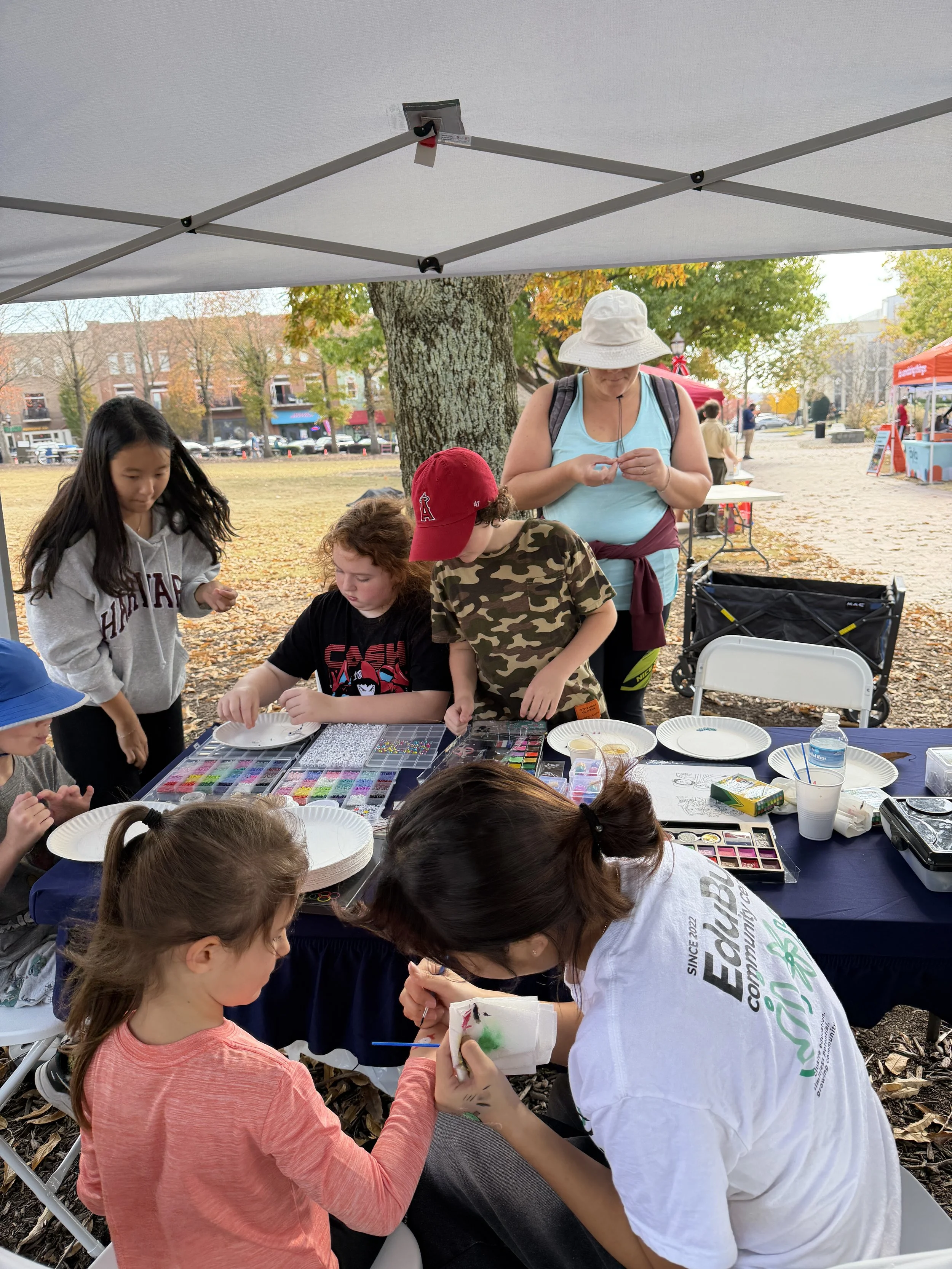 Children and adults at an outdoor craft activity table, under a canopy, with items like beads, paints, and paper, with trees and tents in the background during fall.