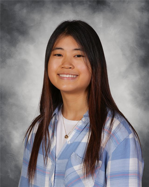A young woman with long dark hair wearing a blue plaid shirt and a necklace, smiling against a gray cloudy background.