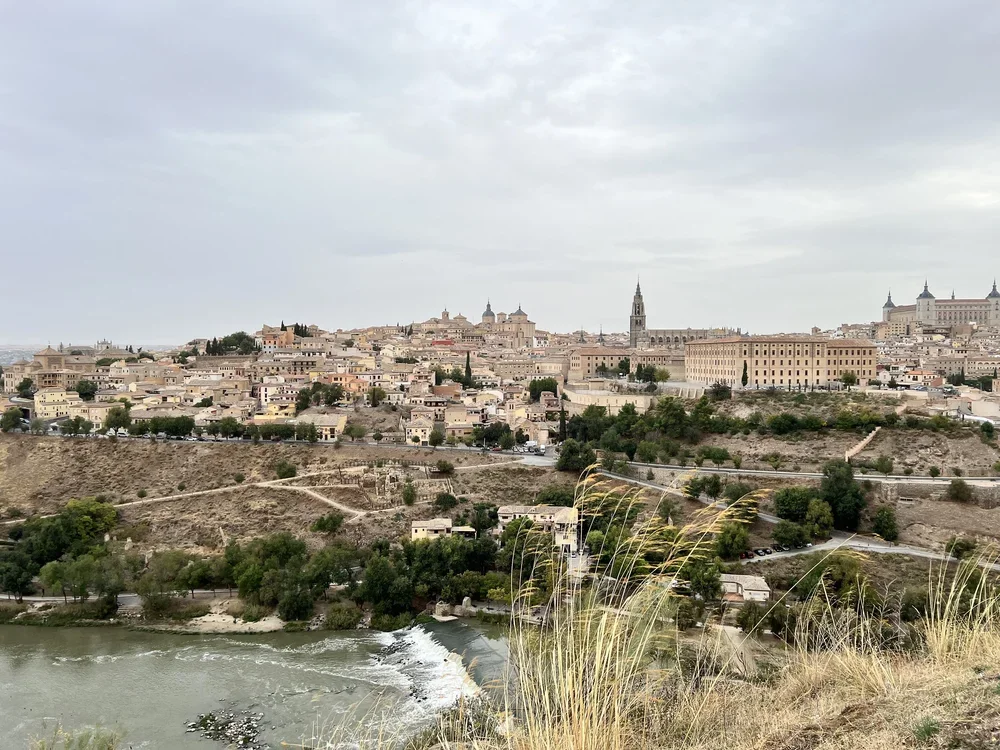 A cityscape of historic buildings on a hill with a river in the foreground, under a cloudy sky.