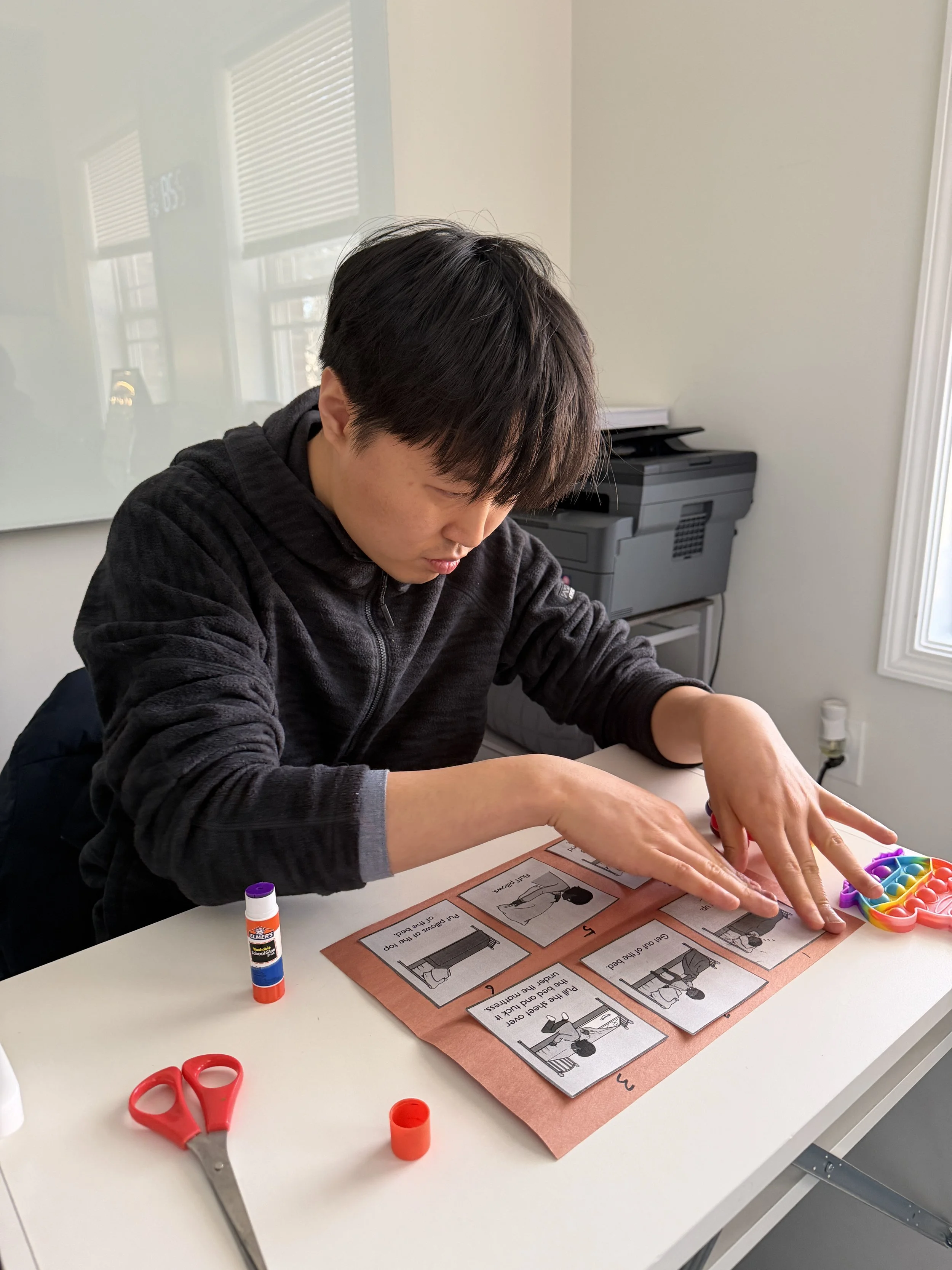 A person working on a visual activity at a desk, with various craft supplies such as scissors, glue, and small colorful items. The activity sheet on the desk contains illustrations and instructions.