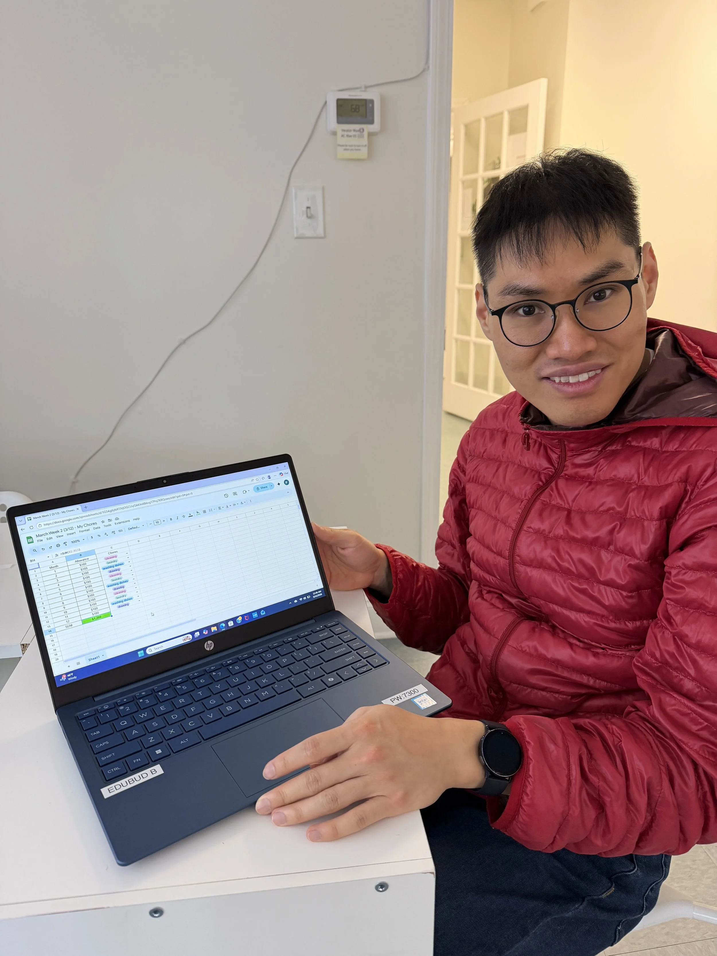 A man with glasses and a black watch, wearing a red jacket, is sitting at a white desk, holding a laptop with a spreadsheet on the screen. The background shows a beige wall, a doorway with glass panels, and a thermostat on the wall.
