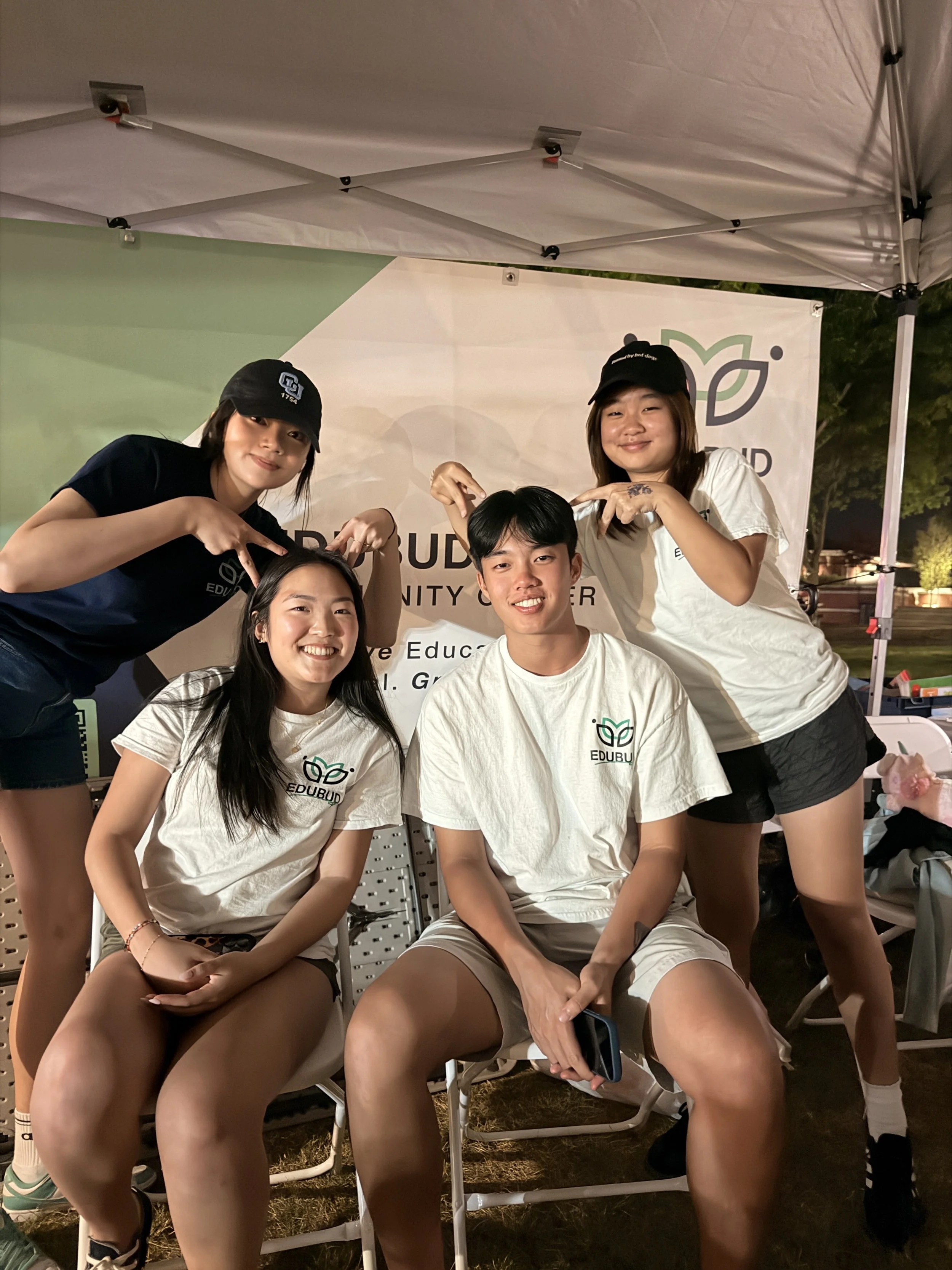 Group of young people, three women, and one man, sitting and standing together at an outdoor event under a canopy tent, smiling and posing for the photo. They are casually dressed, some wearing hats and t-shirts with a leaf logo.