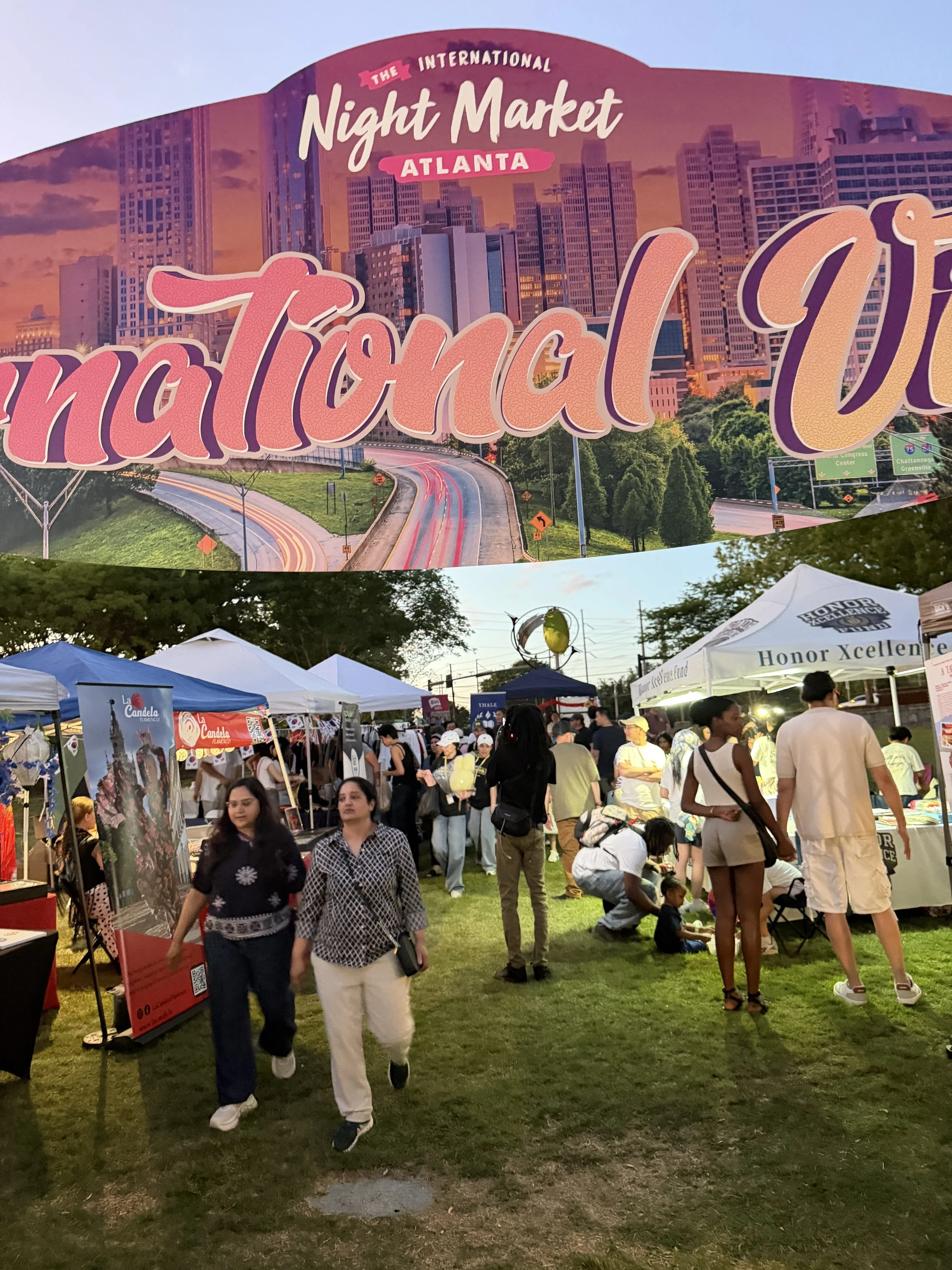 A busy outdoor night market with tents and stalls, many people walking and shopping, with a large city skyline and a sign reading "The International Night Market Atlanta" in the background.