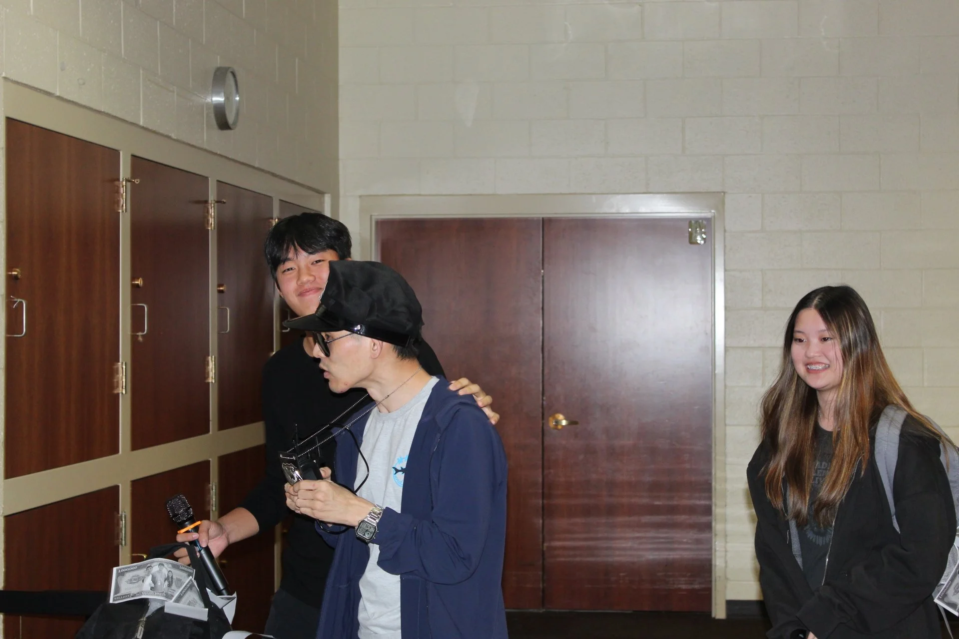 A group of teenagers standing near lockers in a school hallway, one boy with dark hair smiling at the camera, another boy with glasses looking down, and a girl with long hair smiling.