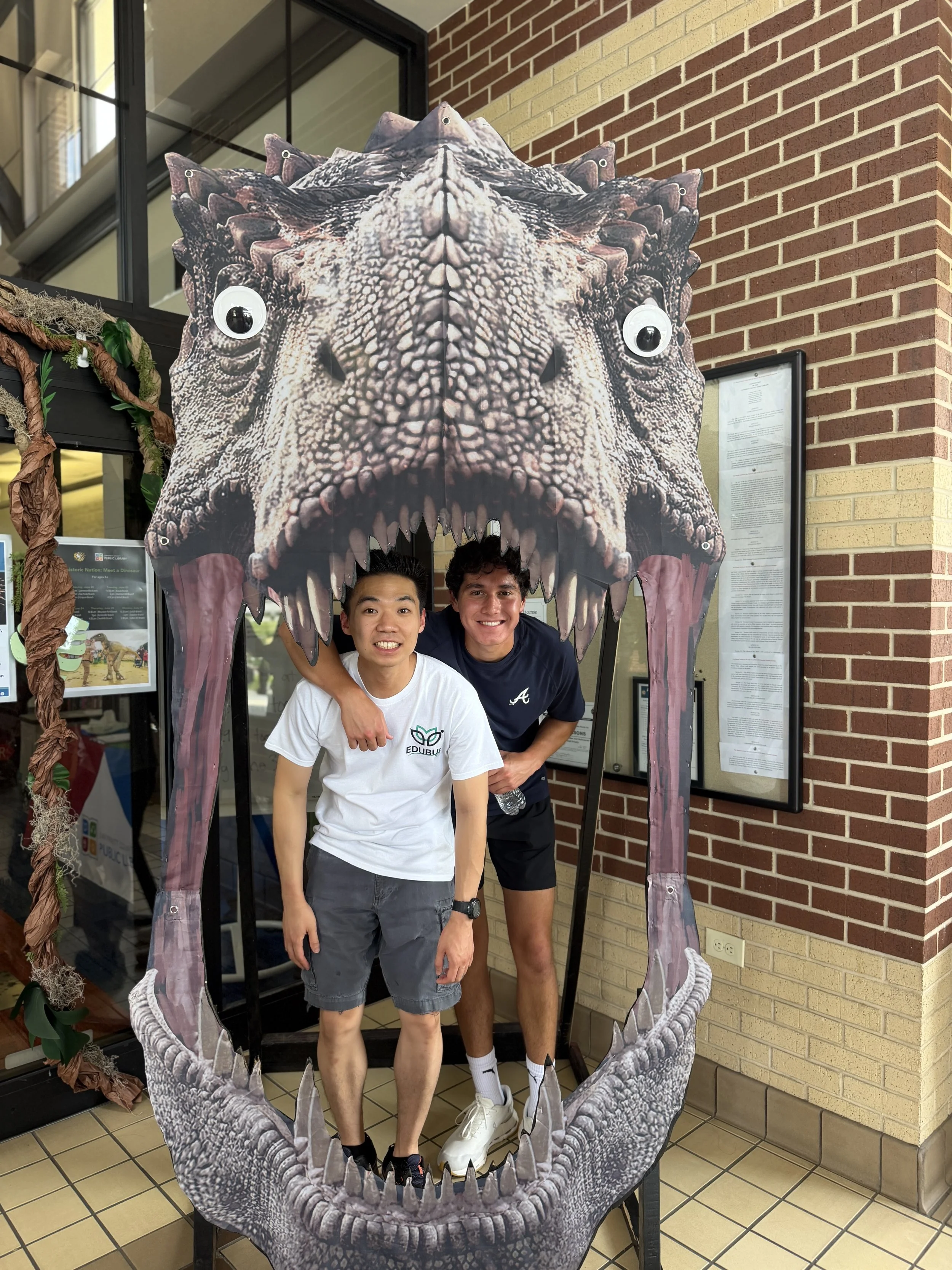 Two young men are posing inside a large dinosaur-themed photo frame with an open mouth, resembling a T-Rex. They are smiling and standing close together.