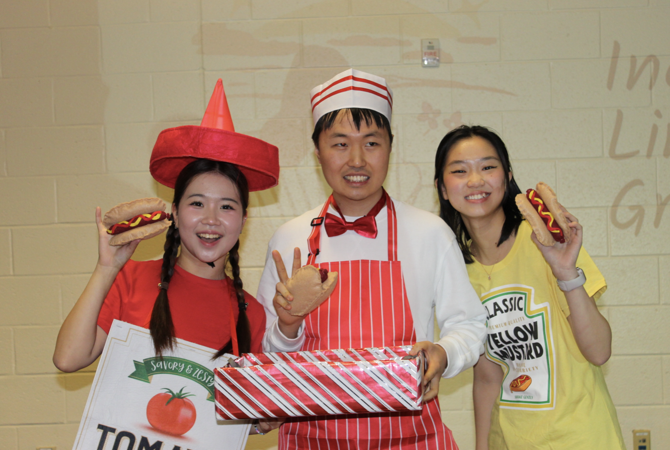 Three people dressed as hot dogs and a chef, smiling and holding hot dog props, standing in front of a light-colored wall.